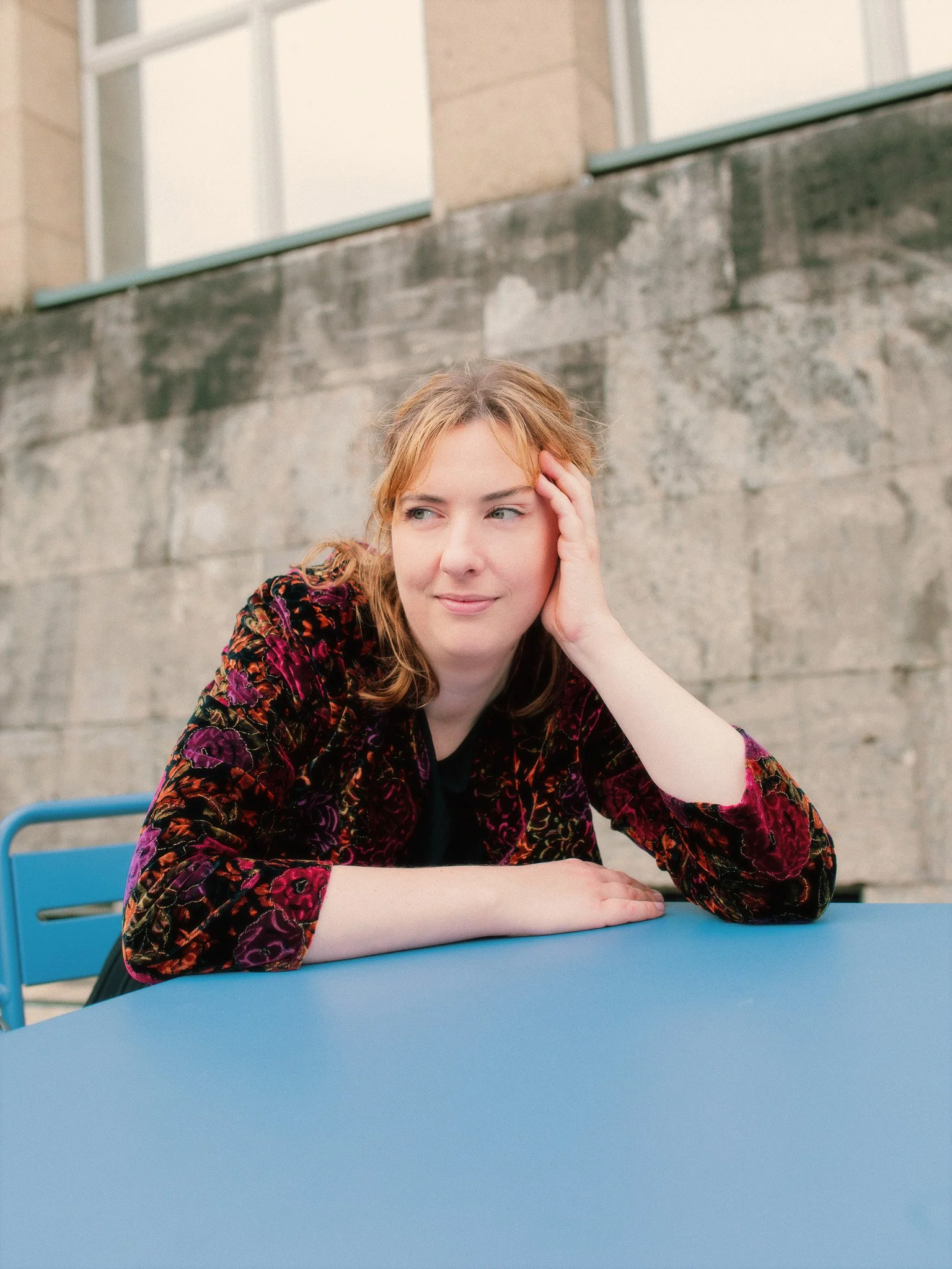 Junge Frau mit rotem Haar und bunter Jacke sitzt an einem blauen Tisch im Freien, hat die Hand an den Kopf, schaut nach rechts, vor einem Betonmauer und einem Gebäude mit Fenstern.