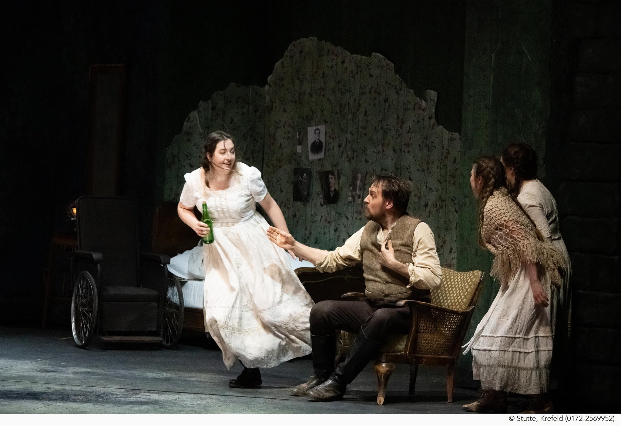 Eine Theateraufführung mit vier Schauspielern in Kostümen. Eine Frau in einem weißen Kleid hält eine Flasche und sitzt in einem Rollstuhl, während sie mit einem Mann spricht, der in einem Hemd und Weste sitzt. Drei weitere Frauen sitzen auf Stühlen und hören zu. Der Hintergrund ist dunkel mit einem stilisierten Felsen und Fotos an der Wand.