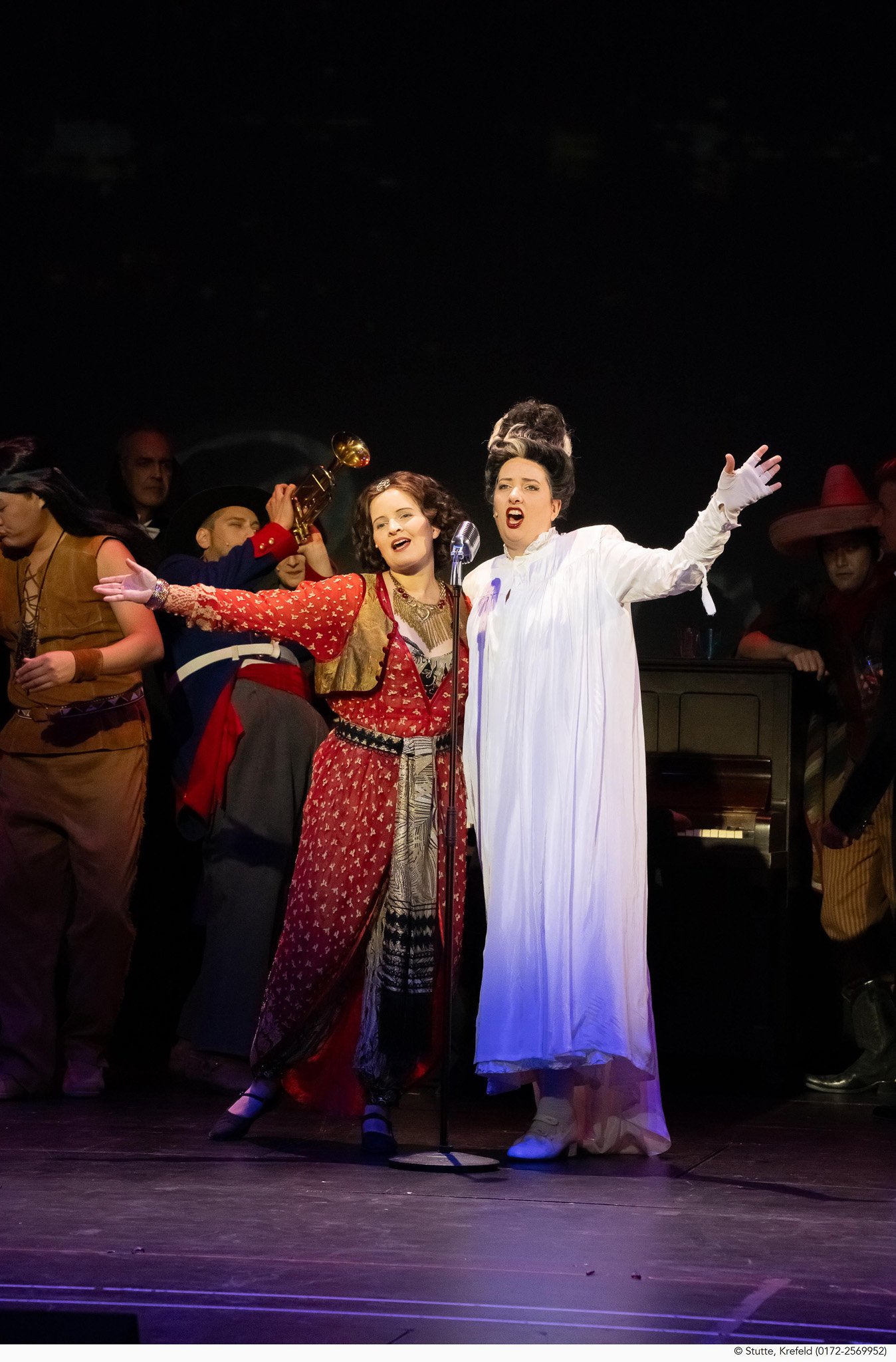 Bühnenszene mit mehreren Schauspielern in historischen Kostümen, die eine Oper oder ein Theaterstück aufführen, darunter zwei Frauen, eine singt an einem Mikrophon, die andere gestikuliert dramatisch.