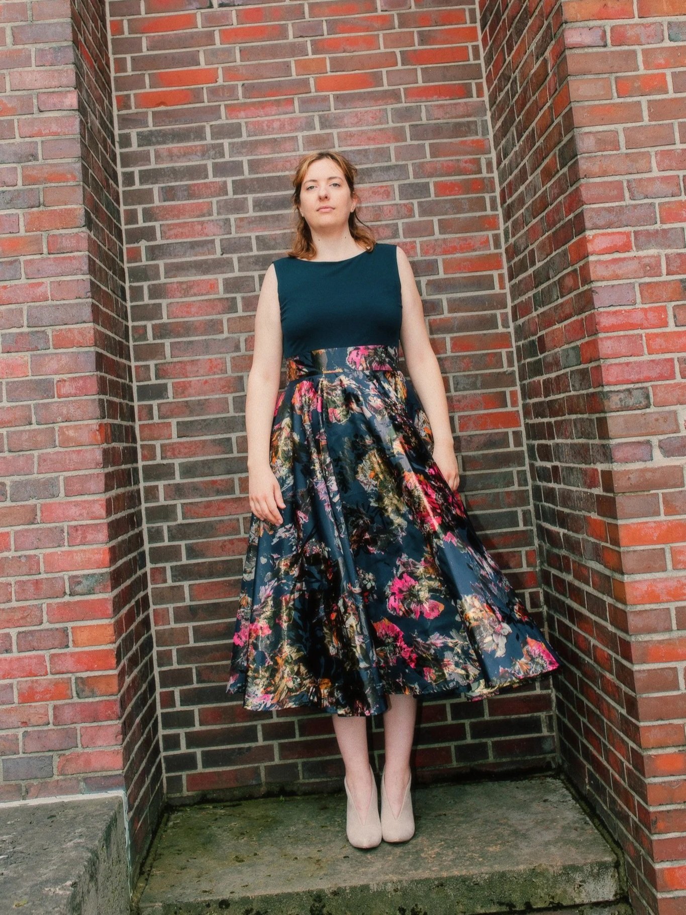 Eine junge Frau steht vor einer roten Backsteinmauer, trägt ein elegantes, ärmelloses dunkelblaues Oberteil und einen langen, farbenfrohen Rock mit Blumenmuster, sowie beige High Heels.