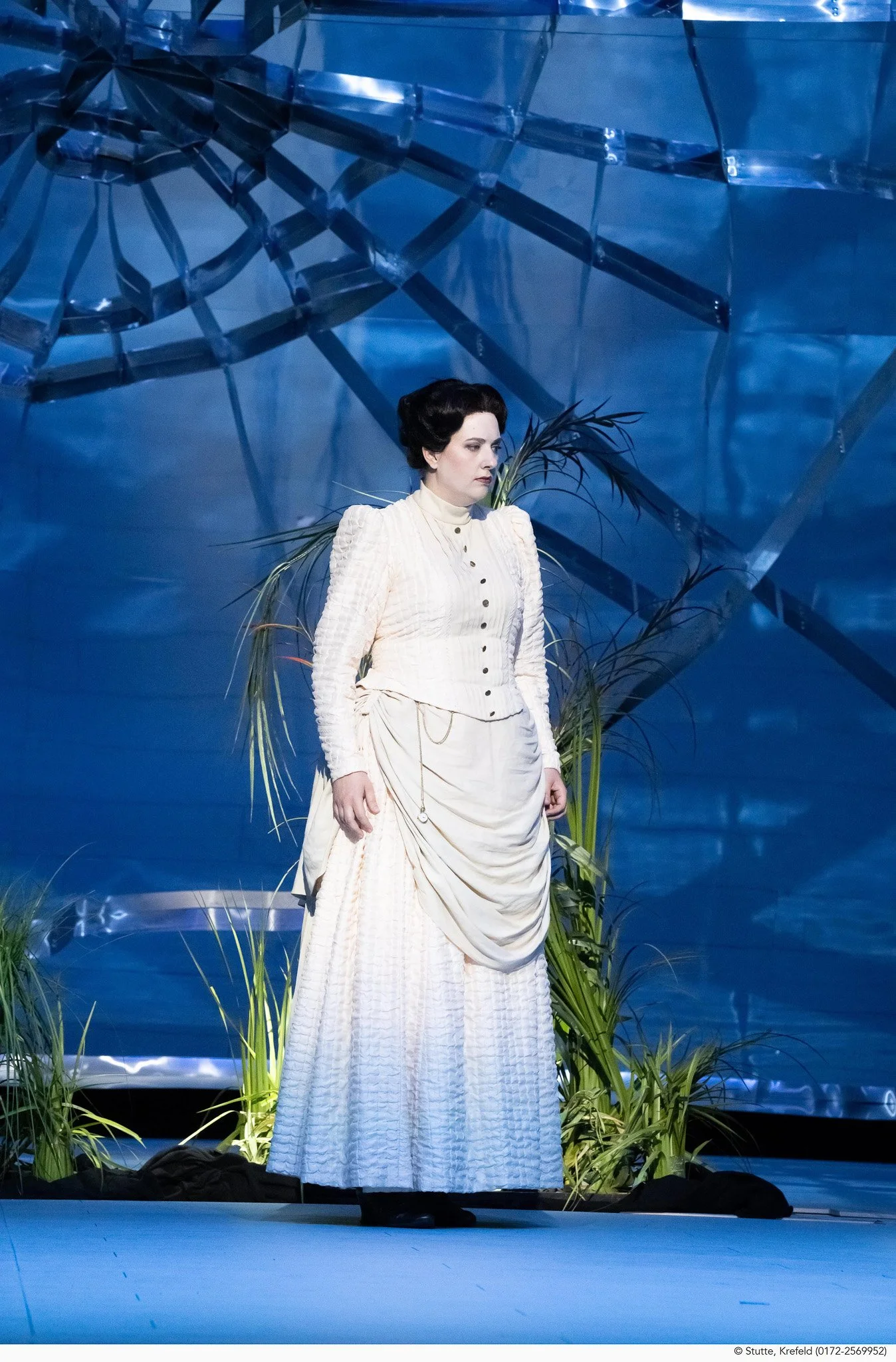 Eine Frau in einem weißen, historischen Kleid steht vor einem blauen Hintergrund mit großen grünen Pflanzen. Sie hat dunkle, hochgesteckte Haare.