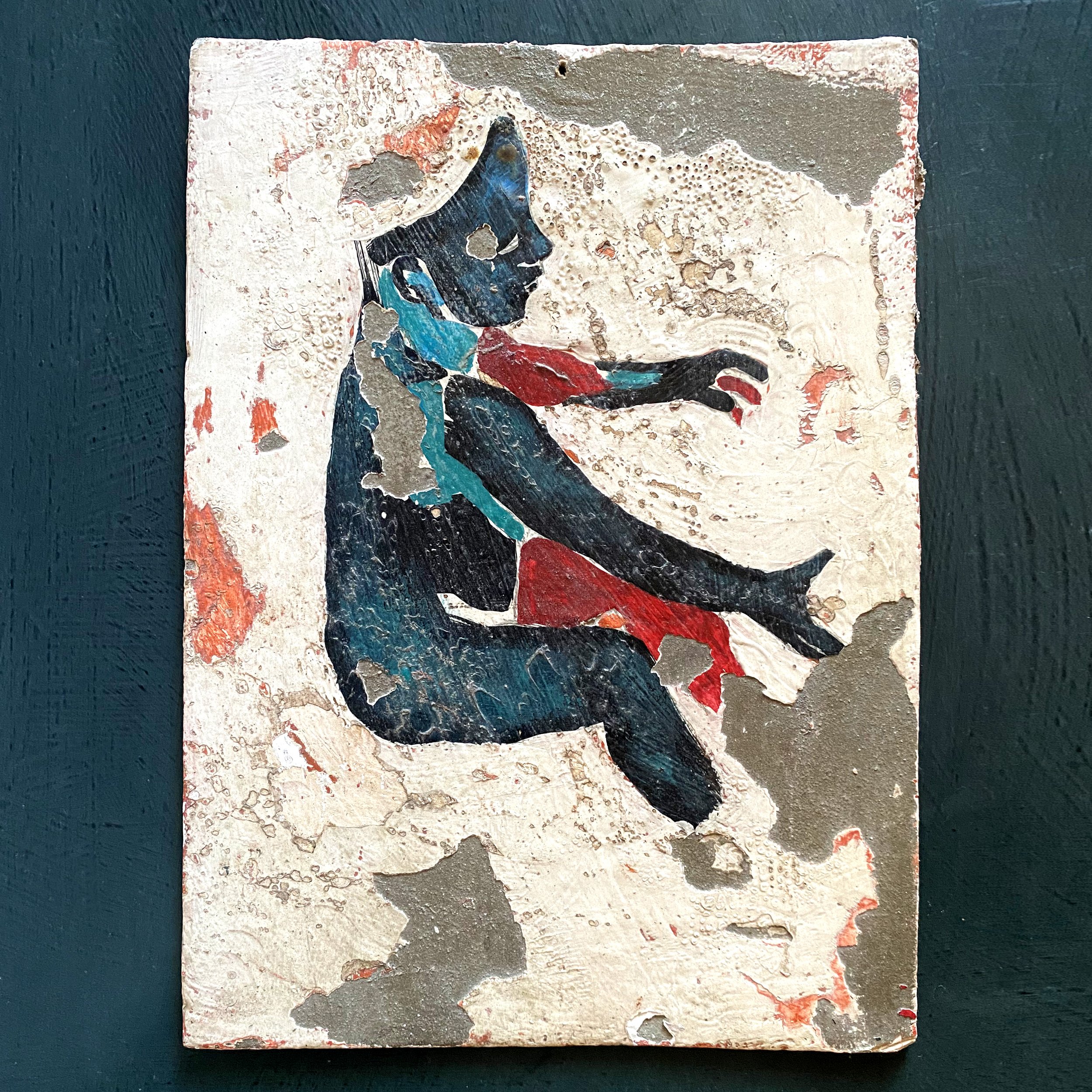 Close-up view of a textured, weathered piece of art or paper with abstract markings, mainly in black, red, and beige, with some blue highlights, on a dark blue background.