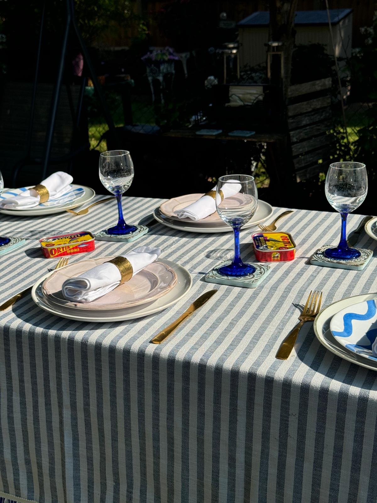 A backyard dining table set for a meal with striped tablecloth, white plates, napkins with gold rings, blue stemware, gold utensils, and small packs of Ortiz anchovies.