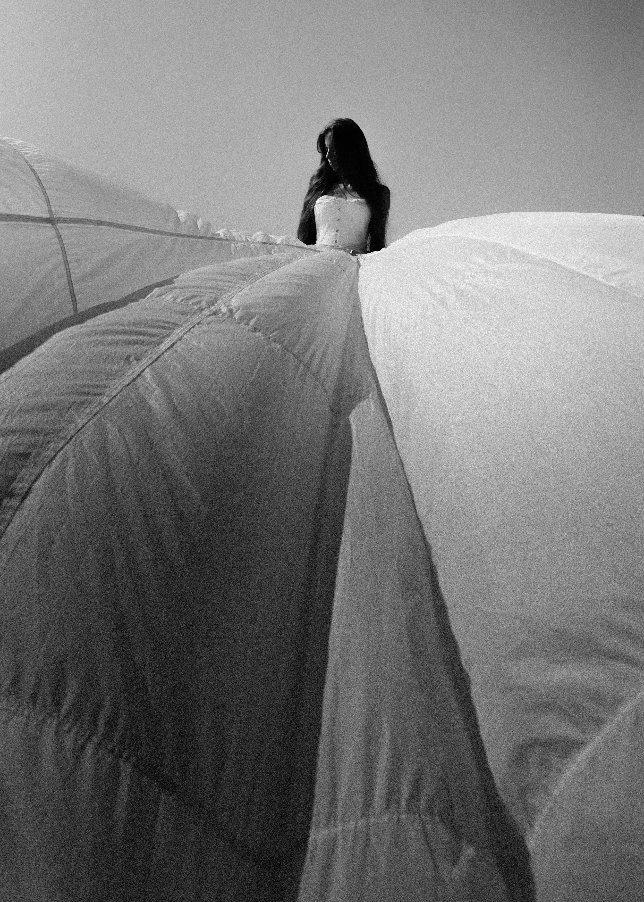 Black and white photo of a woman with long hair standing behind a partially inflated hot air balloon, looking downward.