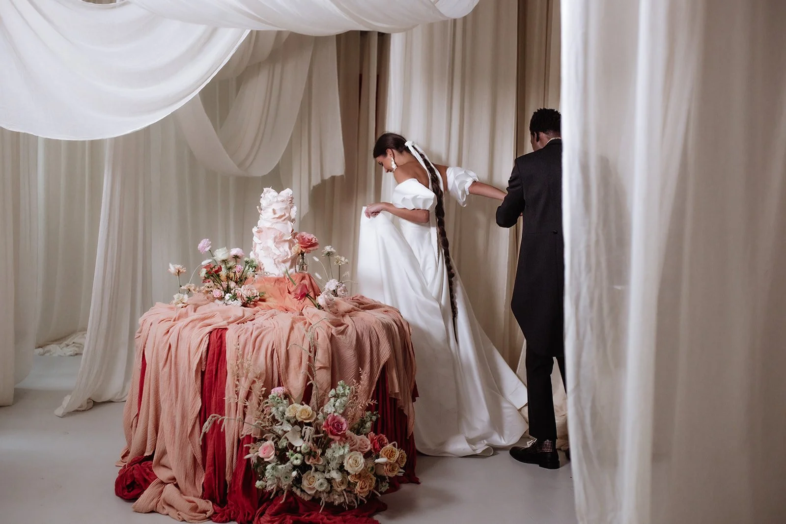 A bride in a white wedding dress and a groom in a black suit standing in a decorated area with off-white drapes, flowers, and a cake.