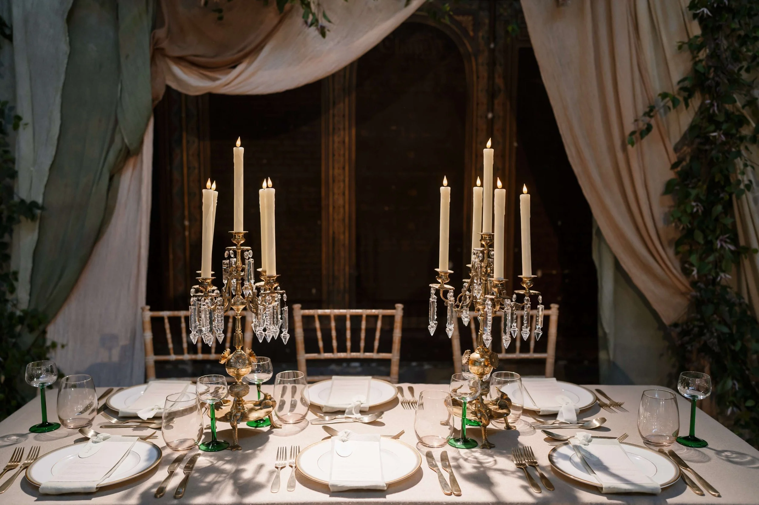 Elegant banquet table set for a formal event with gold and crystal chandelier centerpiece, white plates, silverware, wine glasses, and folded napkins, with draped fabric and greenery in the background.