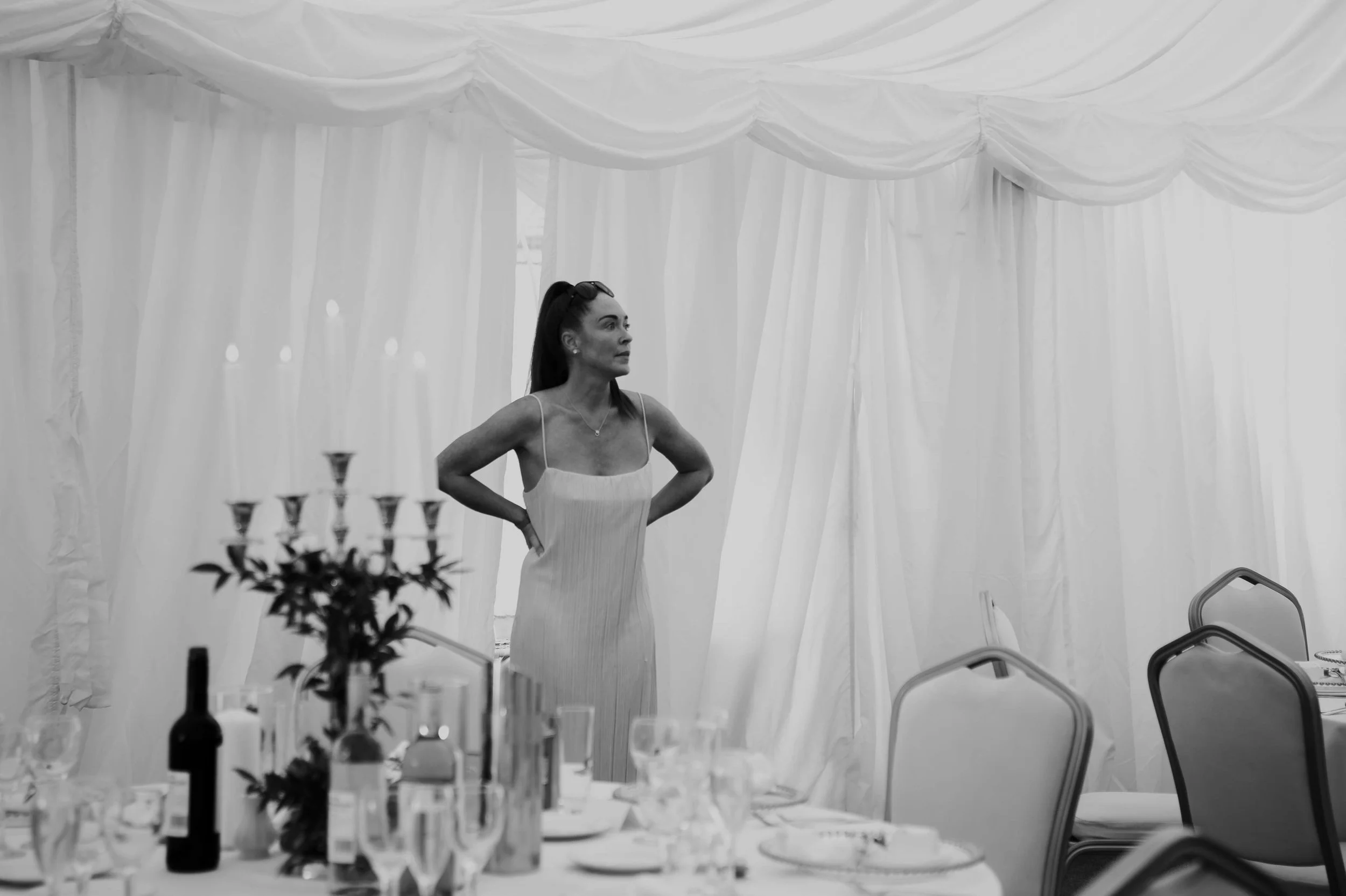 A woman in a strapless dress standing inside a tent with draped white fabric. The table in front of her has wine glasses, bottles, and floral arrangements, suggesting a formal or celebratory event.
