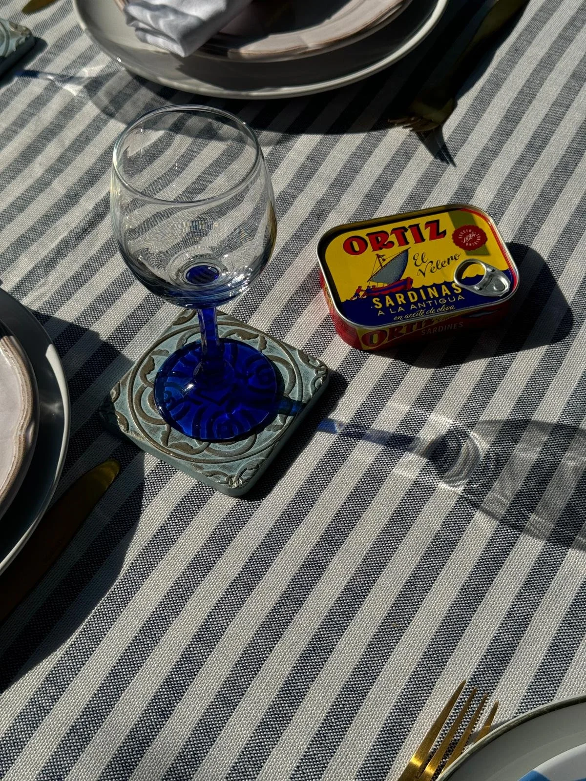 A clear wine glass with a blue stem sitting on a decorative coaster on a table with a striped tablecloth; a tin of Ortiz sardines is nearby.