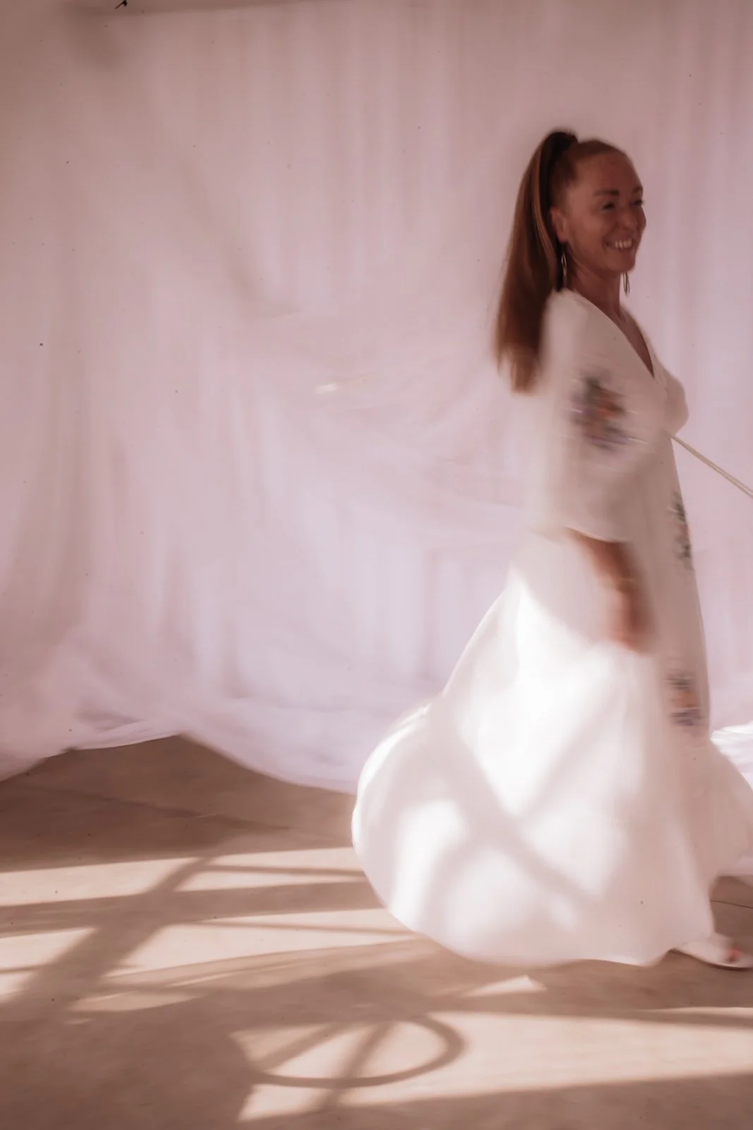 A woman with long hair in a loose white dress spinning and smiling, with a soft pink background and shadows on the floor.