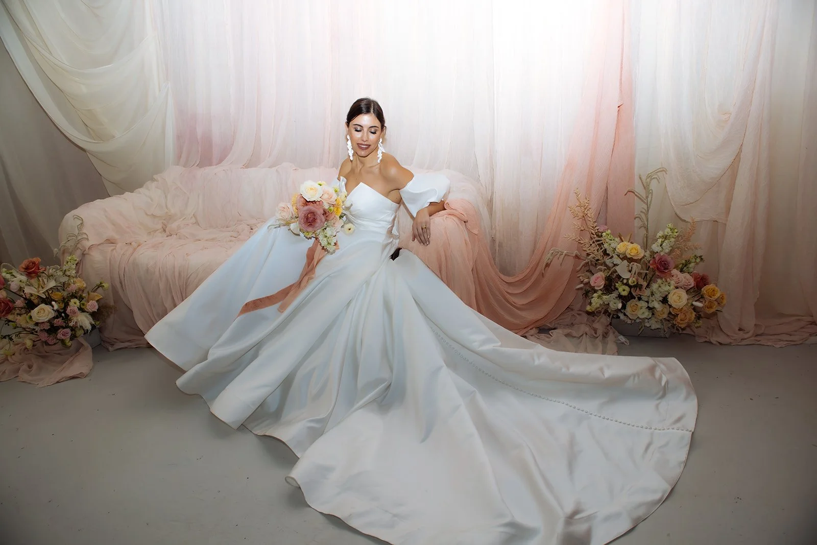 A woman in a white wedding gown sitting on a pink draped sofa, holding a bouquet of flowers, with floral arrangements on either side and soft pastel-colored drapes in the background.