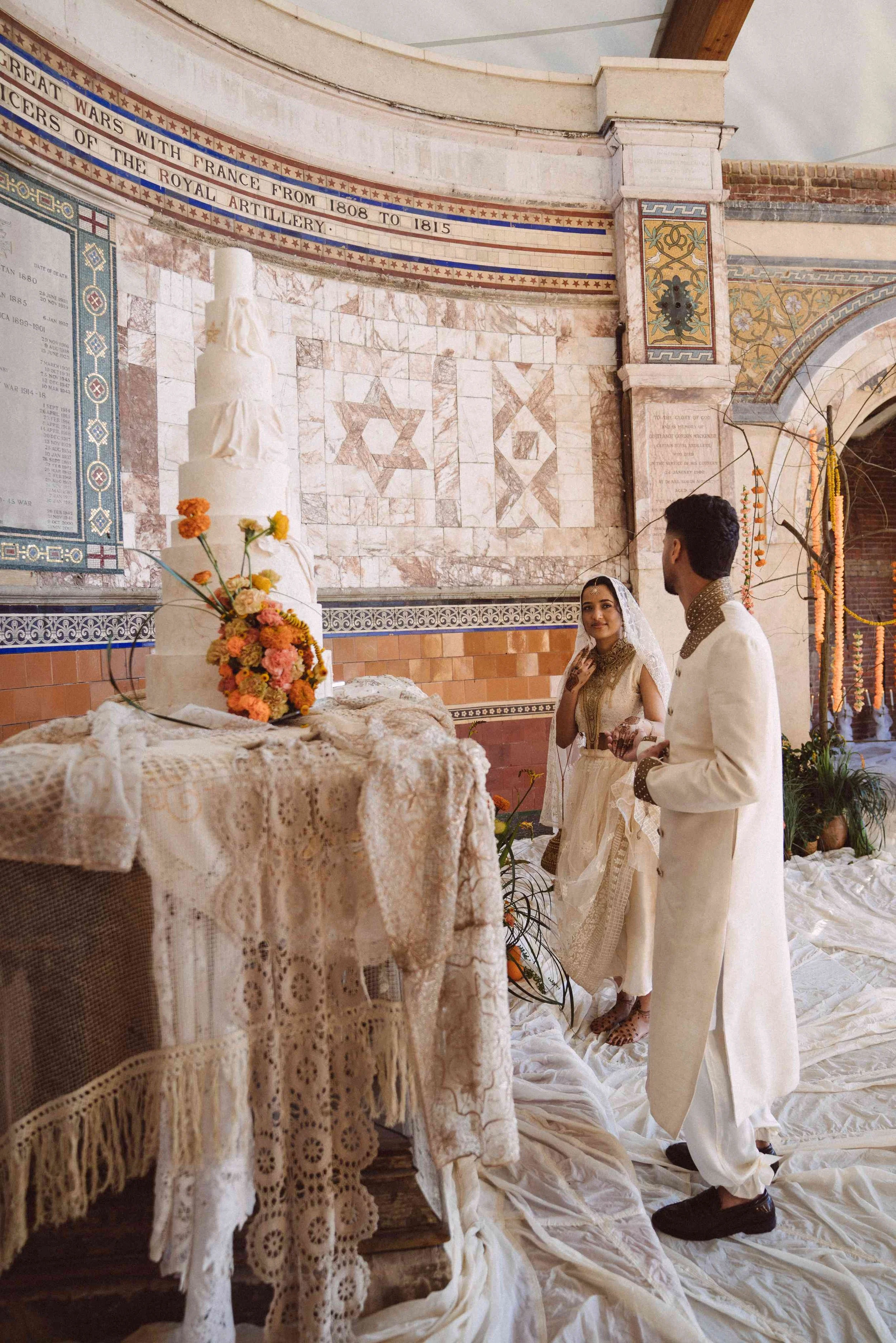 A wedding ceremony inside a church with a couple dressed in traditional Indian wedding attire. The bride is in a cream-colored outfit with a veil, and the groom is in a cream-colored sherwani with black shoes. They are standing near a table with a lace cloth and a floral arrangement. The background features religious symbols, marble decorations, and a wall with historical inscriptions, including one about wars with France.