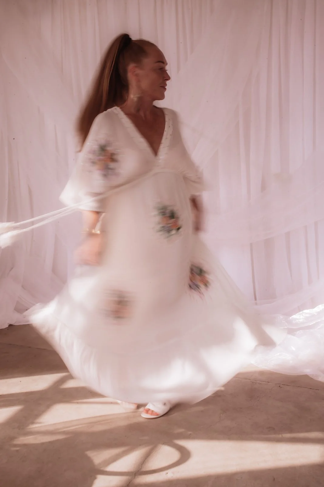A woman in a flowing white dress with floral embroidery twirls around in front of a light-colored fabric backdrop.