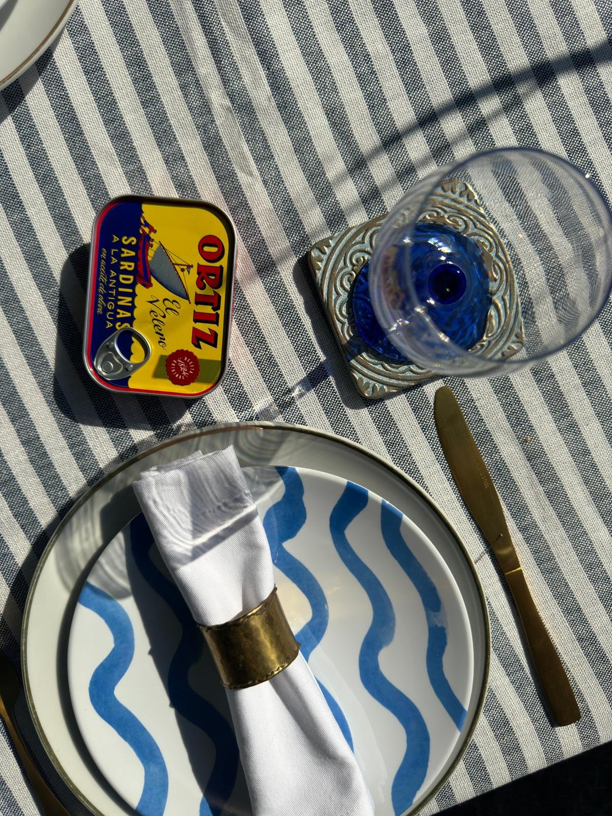 A table setting with a decorative plate holding a white napkin wrapped with a gold ring, a gold-handled knife, a blue and white patterned tablecloth, a small tin of sardines, a clear blue glass, and a white cup or bowl partially visible in the corner.
