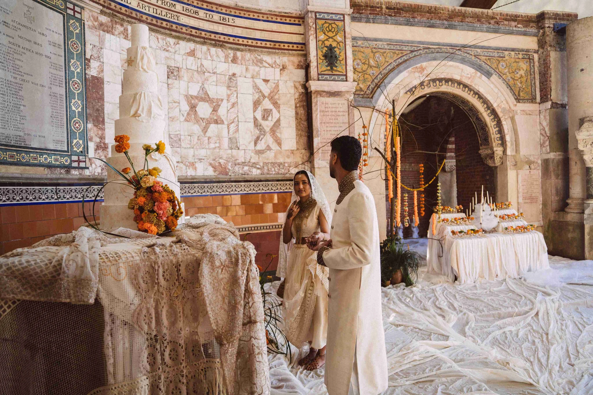 A Hindu wedding ceremony taking place inside a historic temple with intricate mosaic walls. The bride and groom are dressed in traditional cream-colored attire, with the bride wearing a veil and the groom facing her. An altar with lace cloth, flowers, and religious items is visible in the foreground. The background includes a temple arch, and the setting is decorated with marigold garlands and ceremonial items.