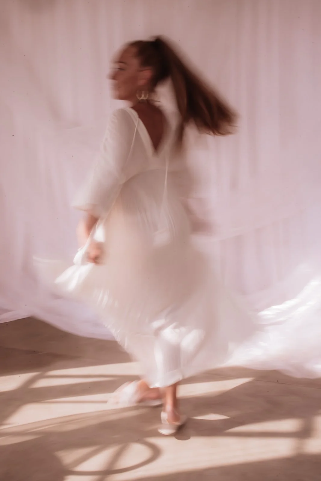 A woman spinning in a white dress, creating a motion blur effect, against a light pink background.