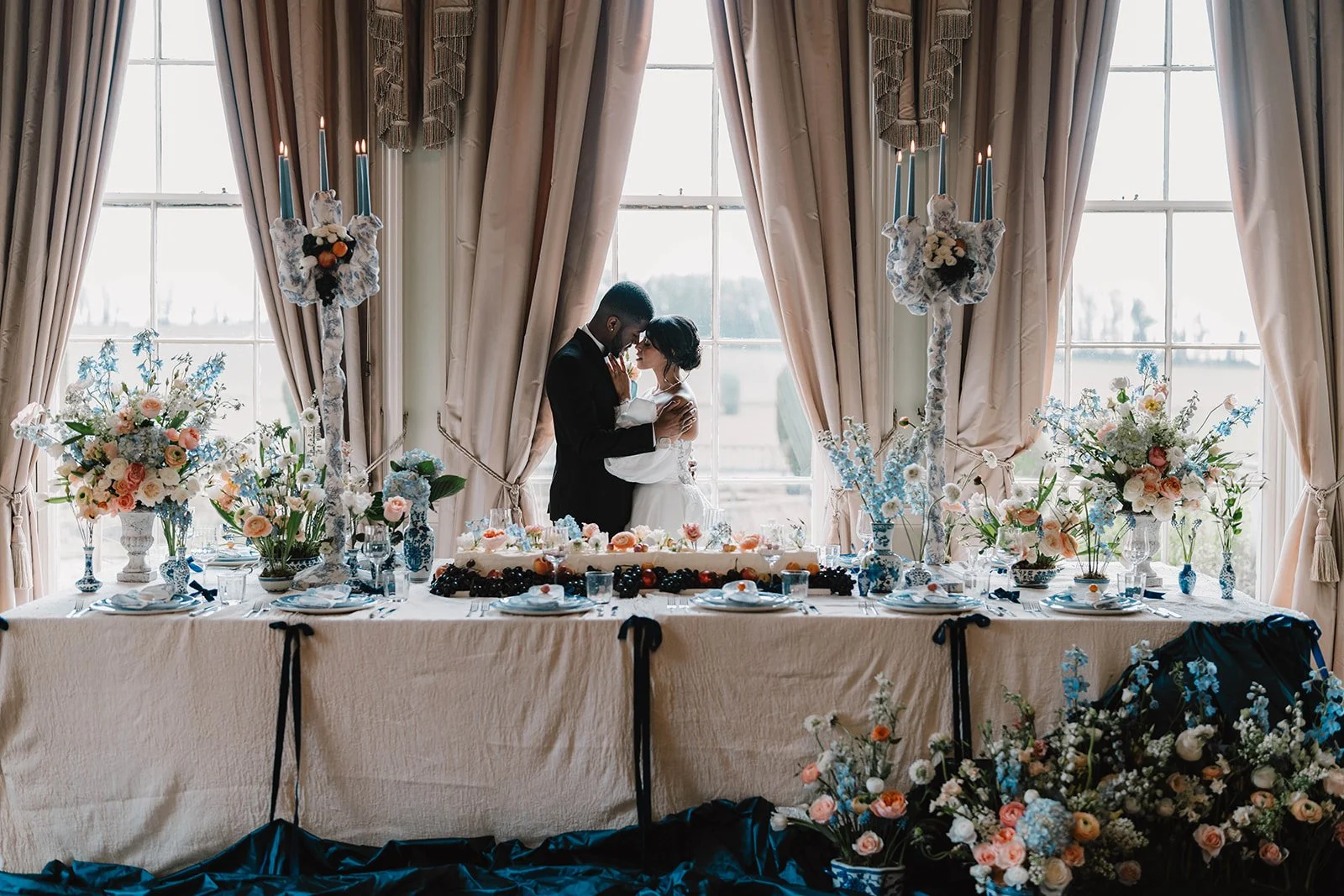 A bride and groom embracing in a wedding reception hall with large windows, decorated with floral arrangements, candles, and a decorated table with a wedding cake.