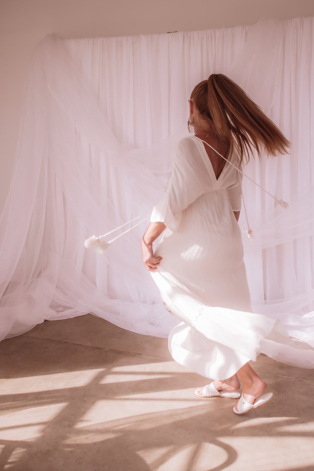 A woman with red hair in a ponytail spinning in a white dress, with sheer white fabric and drapes behind her, in a softly lit room.