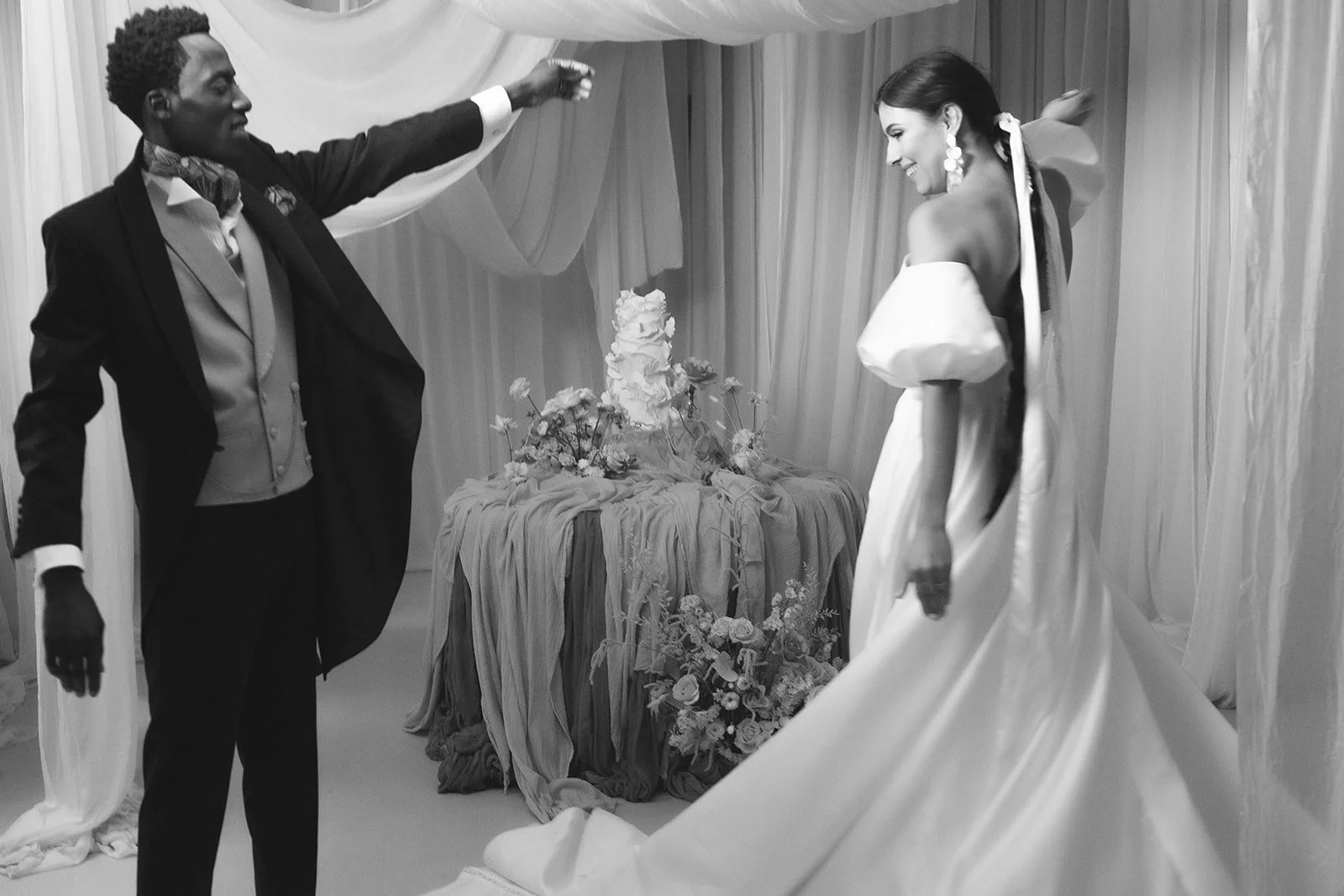 A black and white photo of a couple dancing at their wedding reception. The groom is wearing a tuxedo with a bow tie, and the bride is dressed in an off-the-shoulder gown with large earrings. They are smiling and looking at each other, with a table of flowers and a wedding cake in the background.