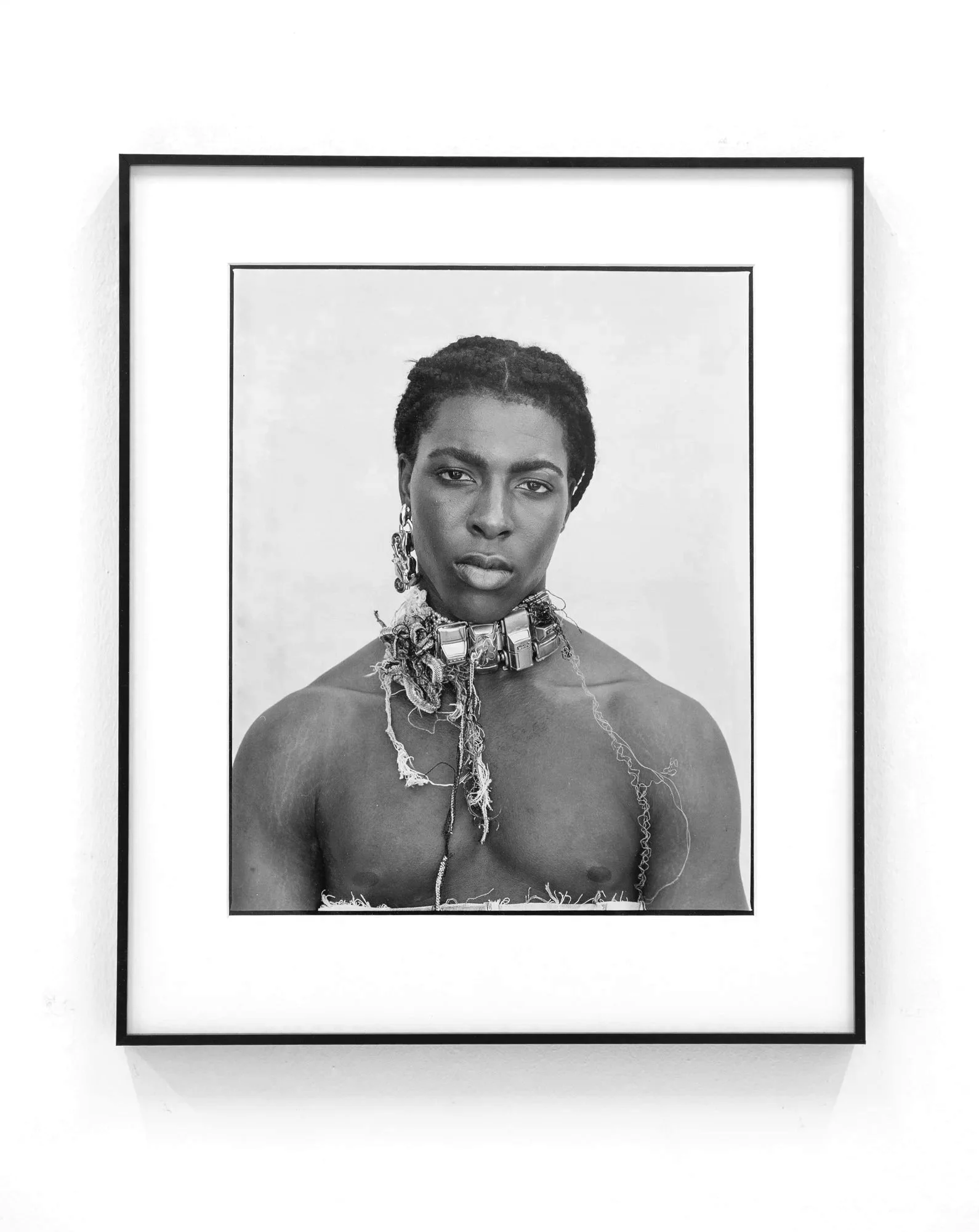 Black and white portrait photograph of a woman with dark hair, styled in short dreadlocks, wearing large hoop earrings, an ornate choker necklace, and reveling in an art exhibit displayed in a black frame mounted on a white wall.