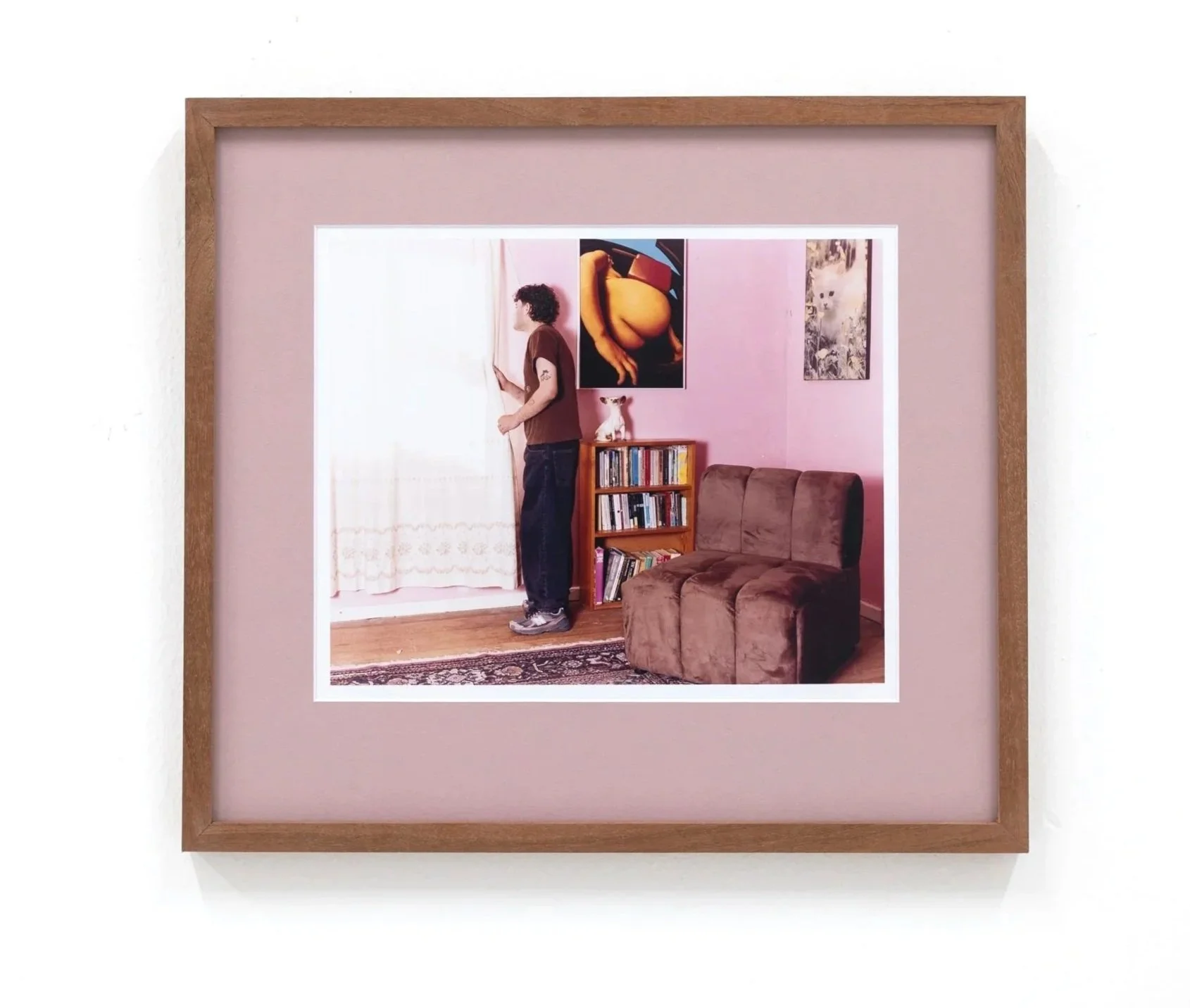 Framed photograph showing a woman with curly hair opening white curtains in a pink-walled room, with artwork, a bookshelf, a brown sofa, and a small white dog in the background.