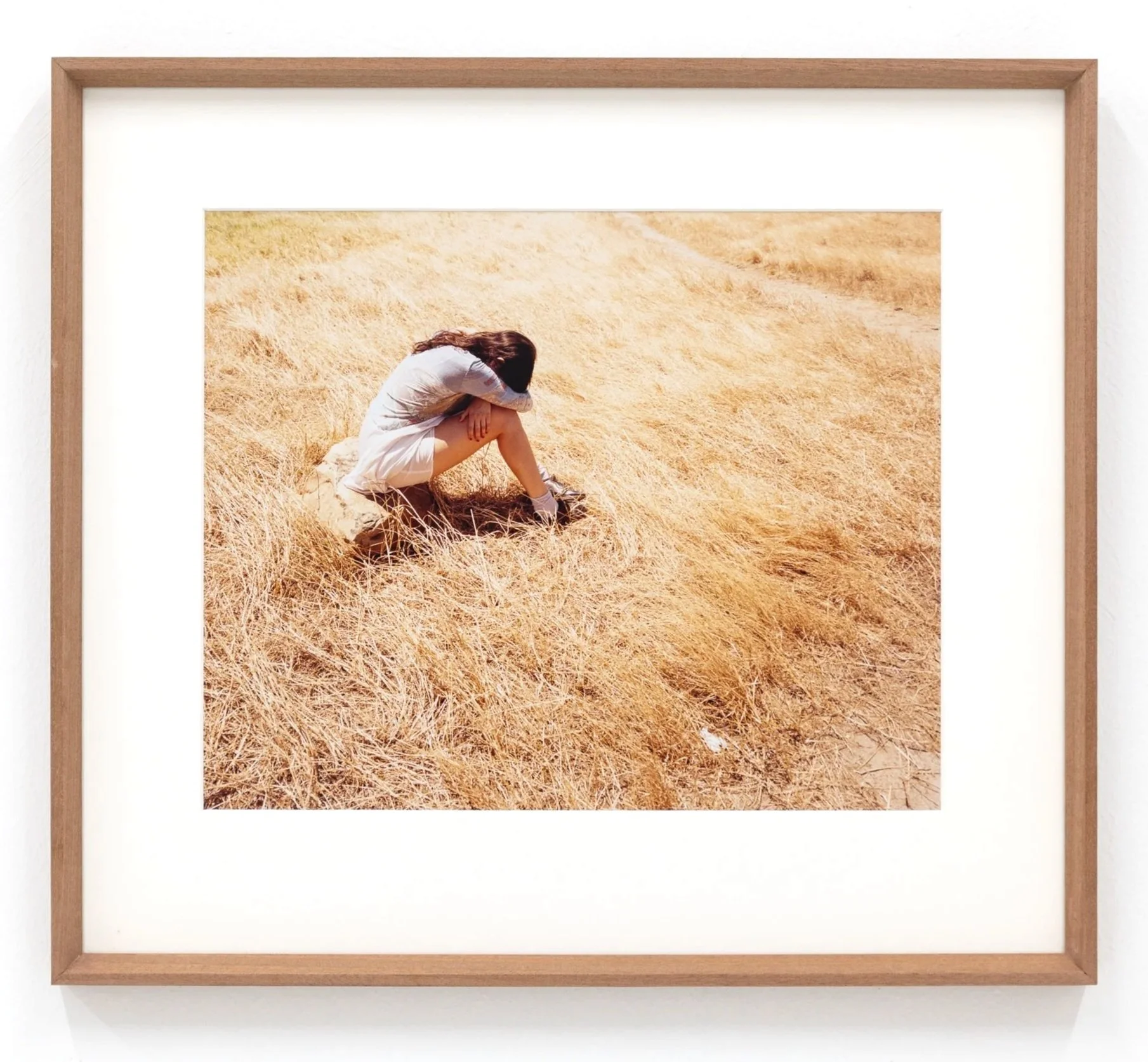 Framed photograph of a woman sitting in a field of tall, dry grass, with her head resting on her knees, wearing a light-colored dress.