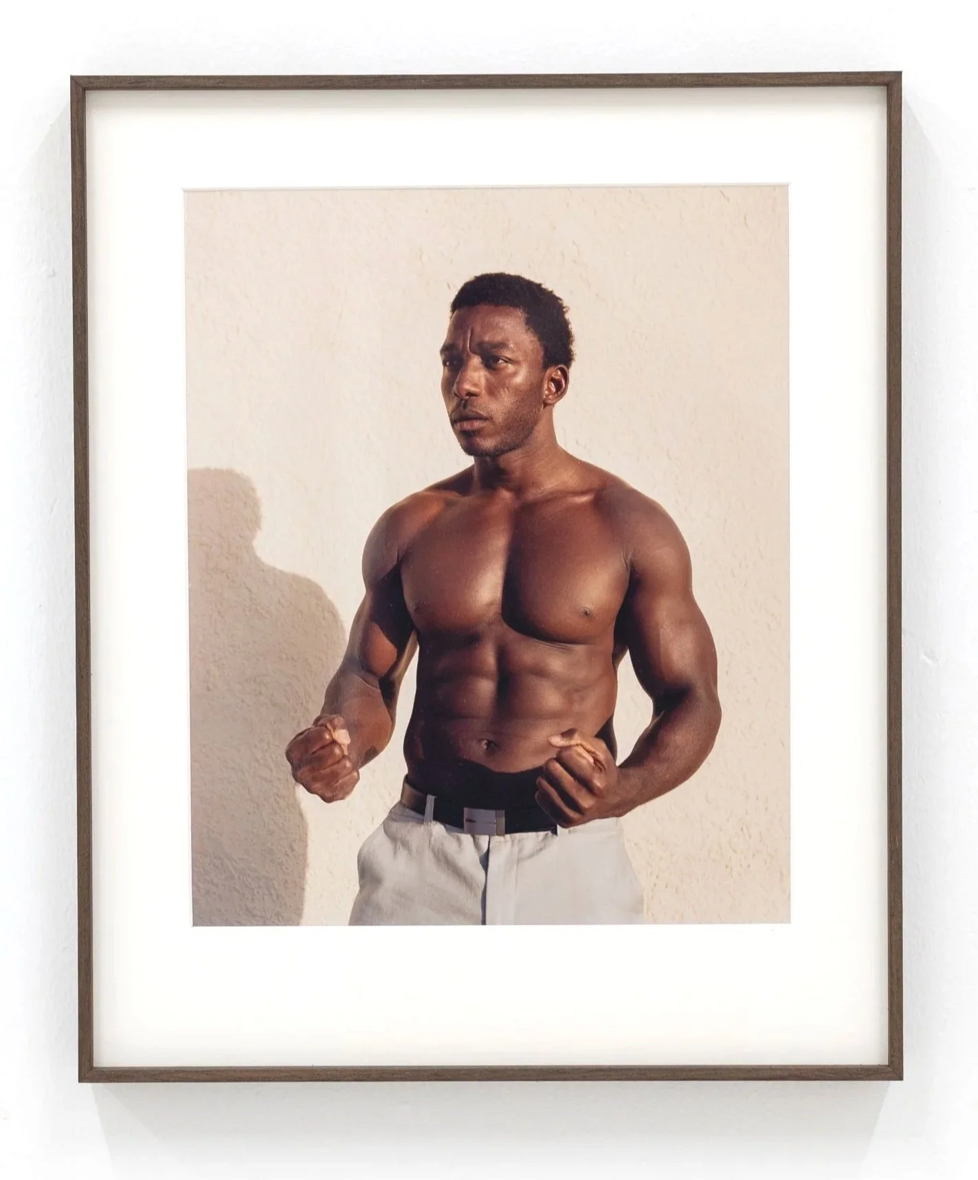 A framed photograph features a shirtless man with a muscular physique, clenched fists, and a serious expression, standing against a plain wall.