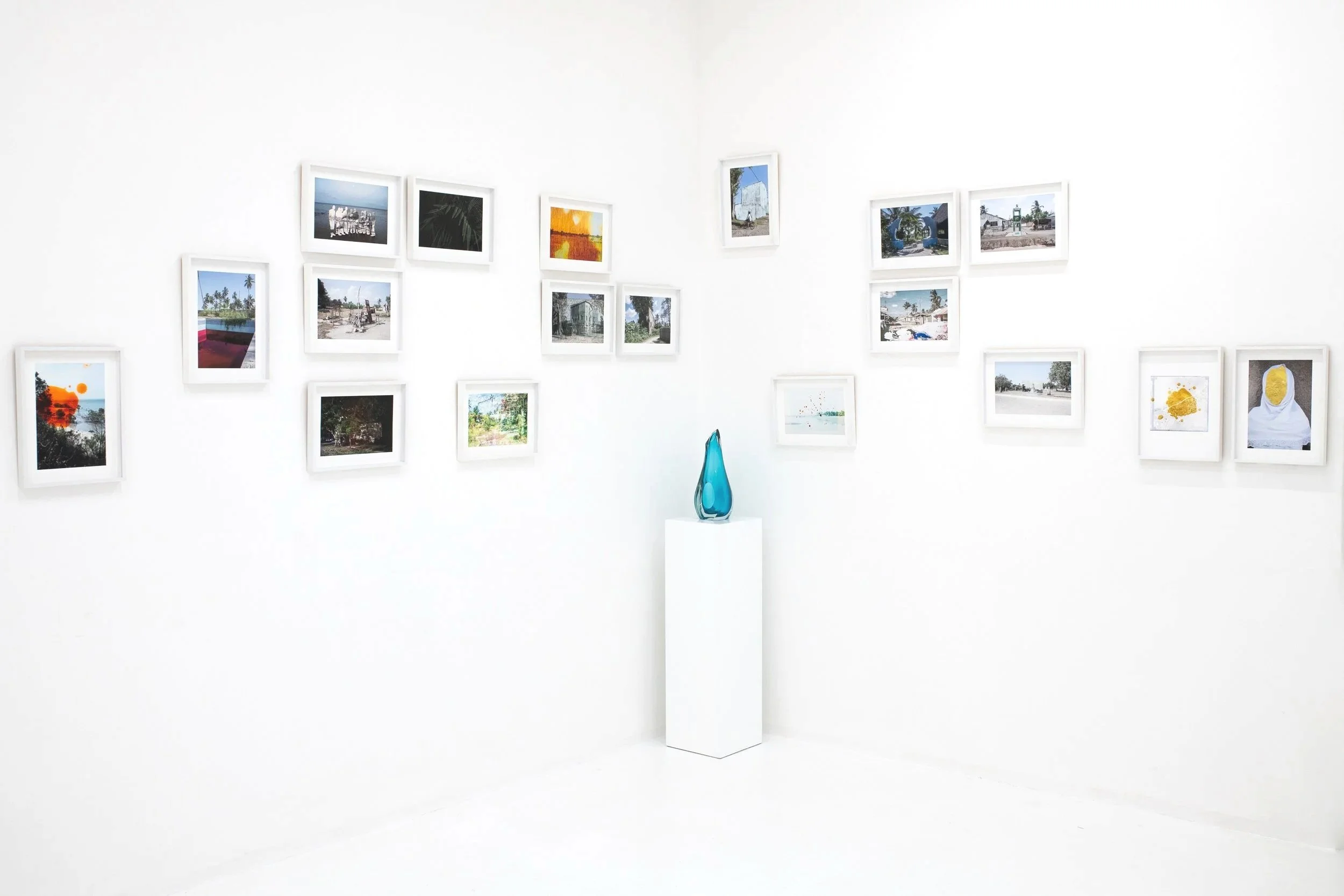 A white art gallery wall with multiple framed photographs and paintings, with a blue glass sculpture on a pedestal in the center.