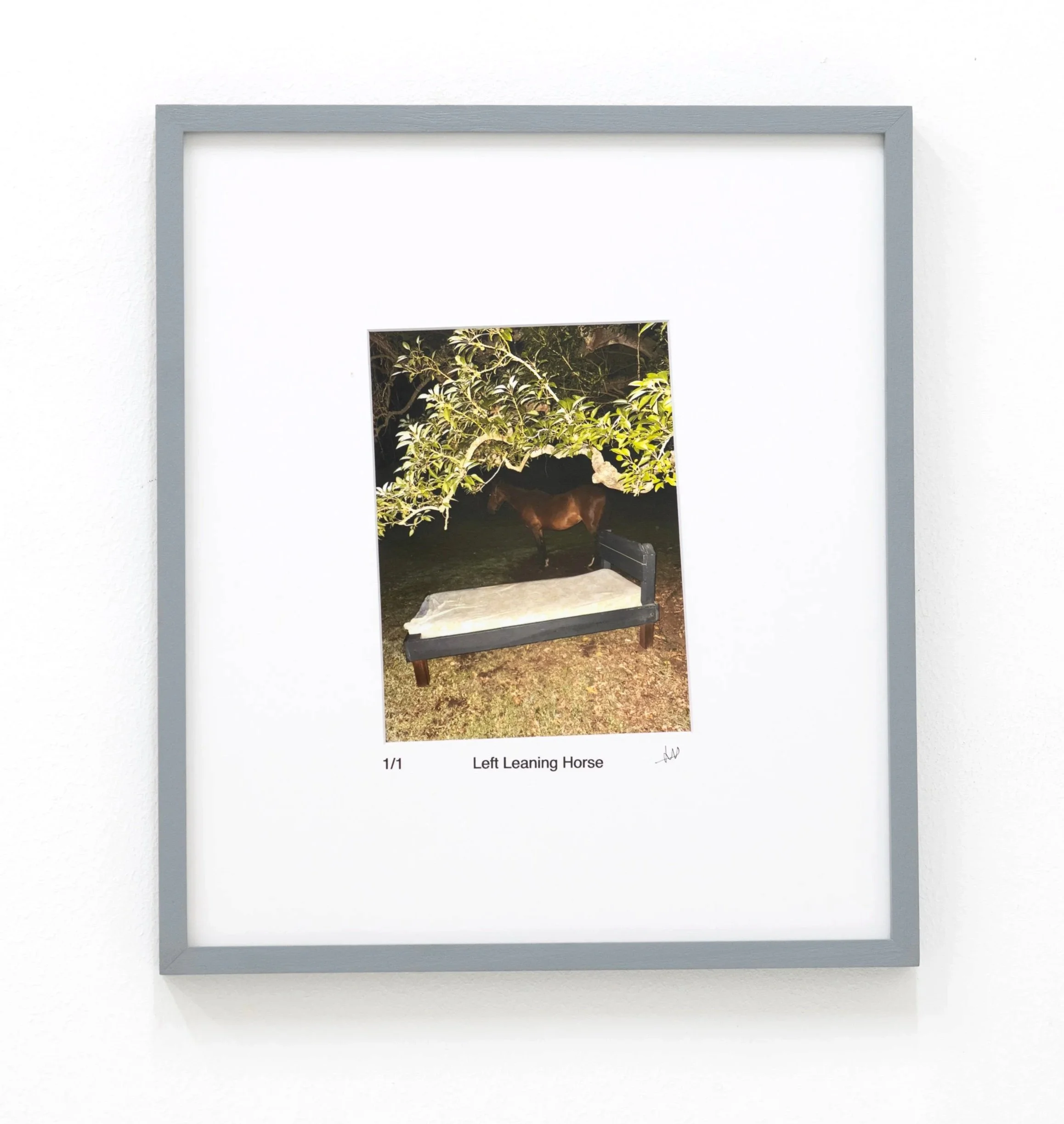 Photograph of a framed artwork titled 'Left Leaning Horse' featuring a nature scene with a brown horse next to trees and bushes, displayed against a white wall.