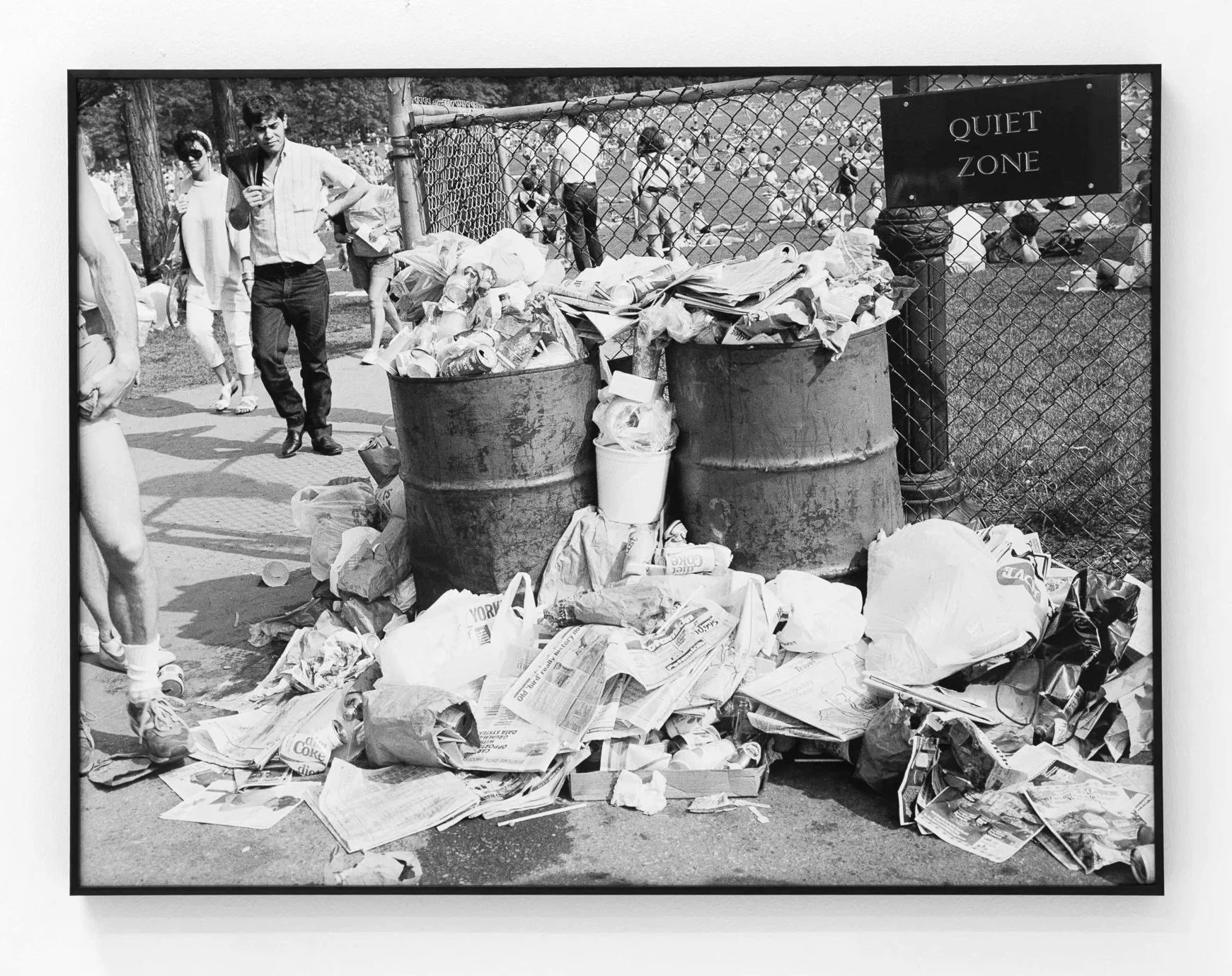 Black and white photograph of a trash bin overflowing with crumpled newspapers and paper waste, placed on a sidewalk near a chain-link fence with a sign that reads 'Quiet Zone'. In the background, people are gathered in a park with trees and sports f