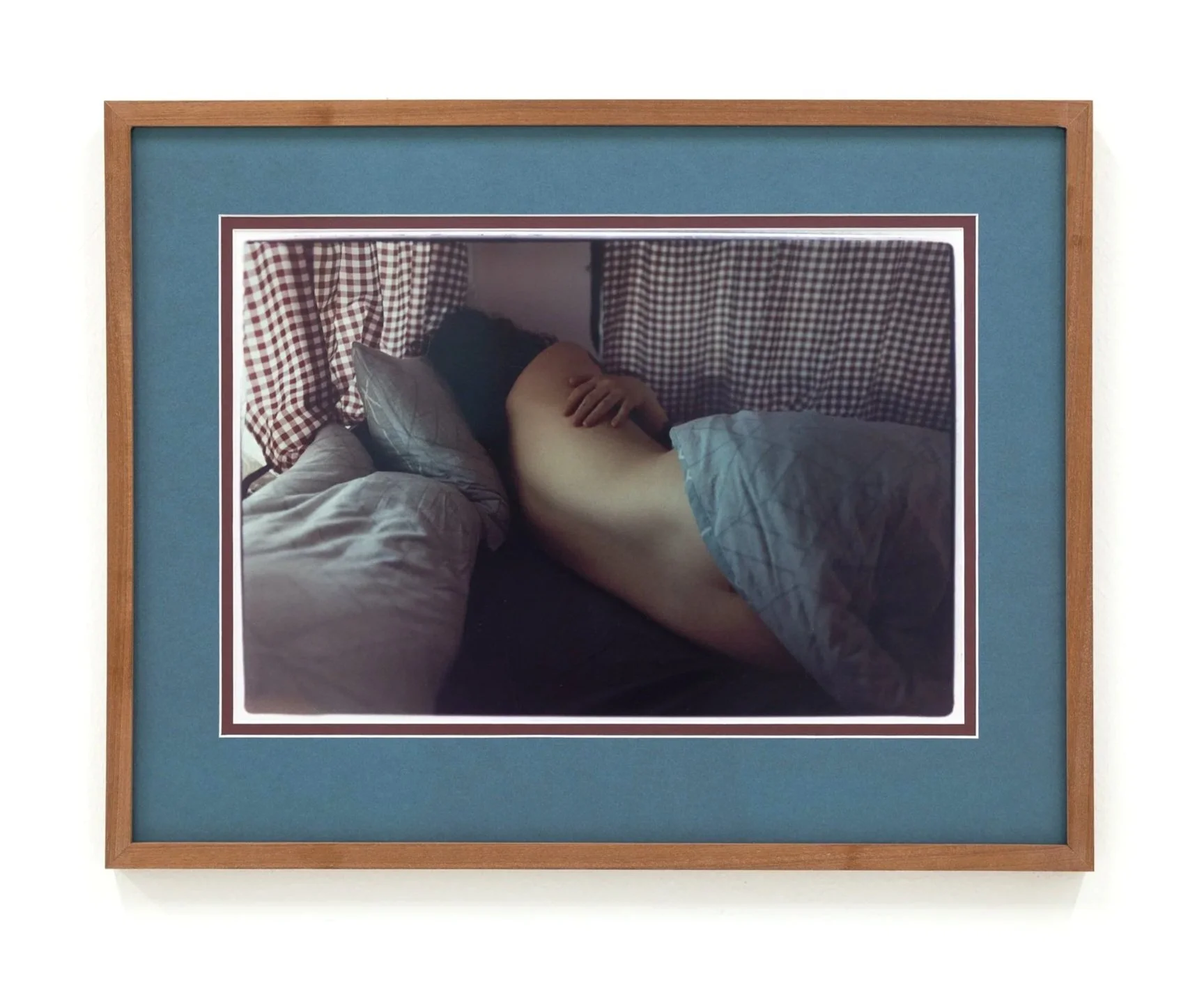 A framed photograph of a person lying in bed, with only their arm and hand visible, resting on their stomach. The bed has a checkered curtain behind it, and the person is under a grey blanket.