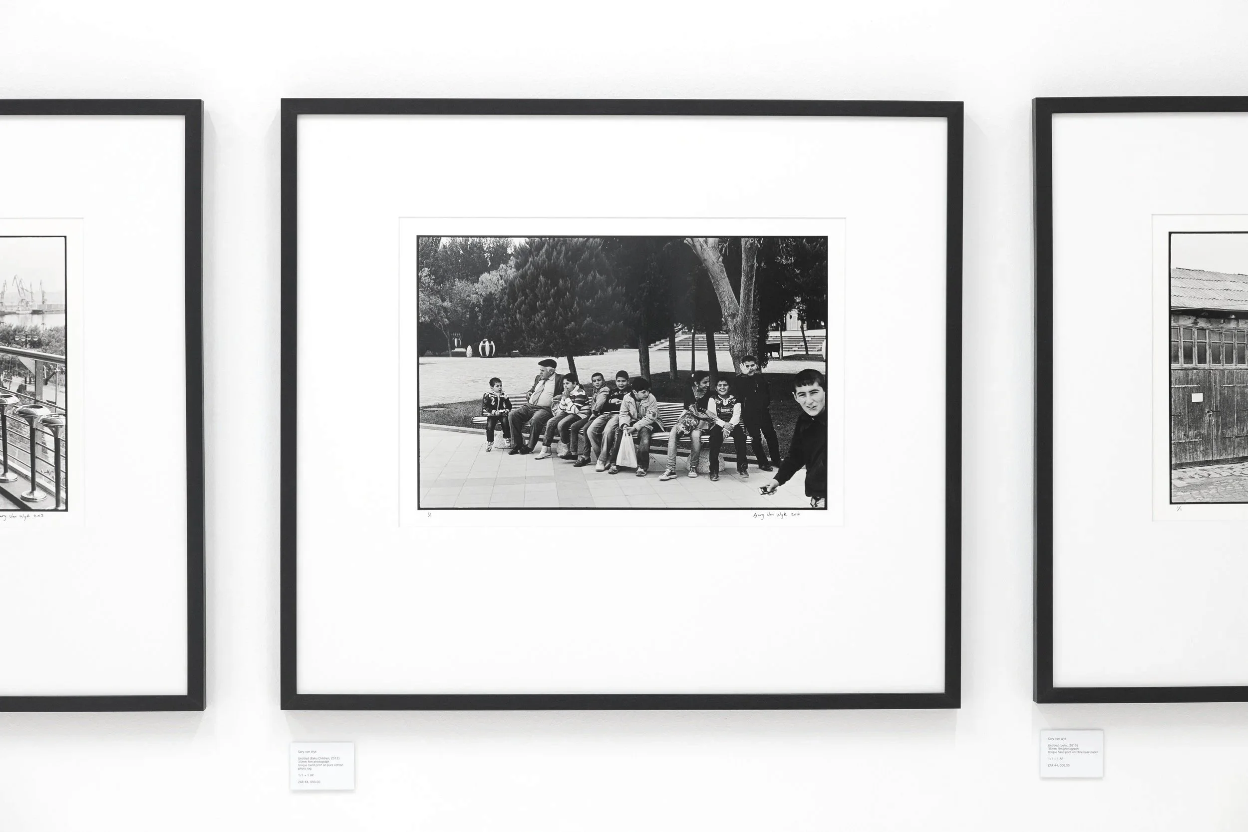 Black and white photograph of a group of children sitting on a park bench with a man, trees, and open space in the background, displayed in an art gallery.