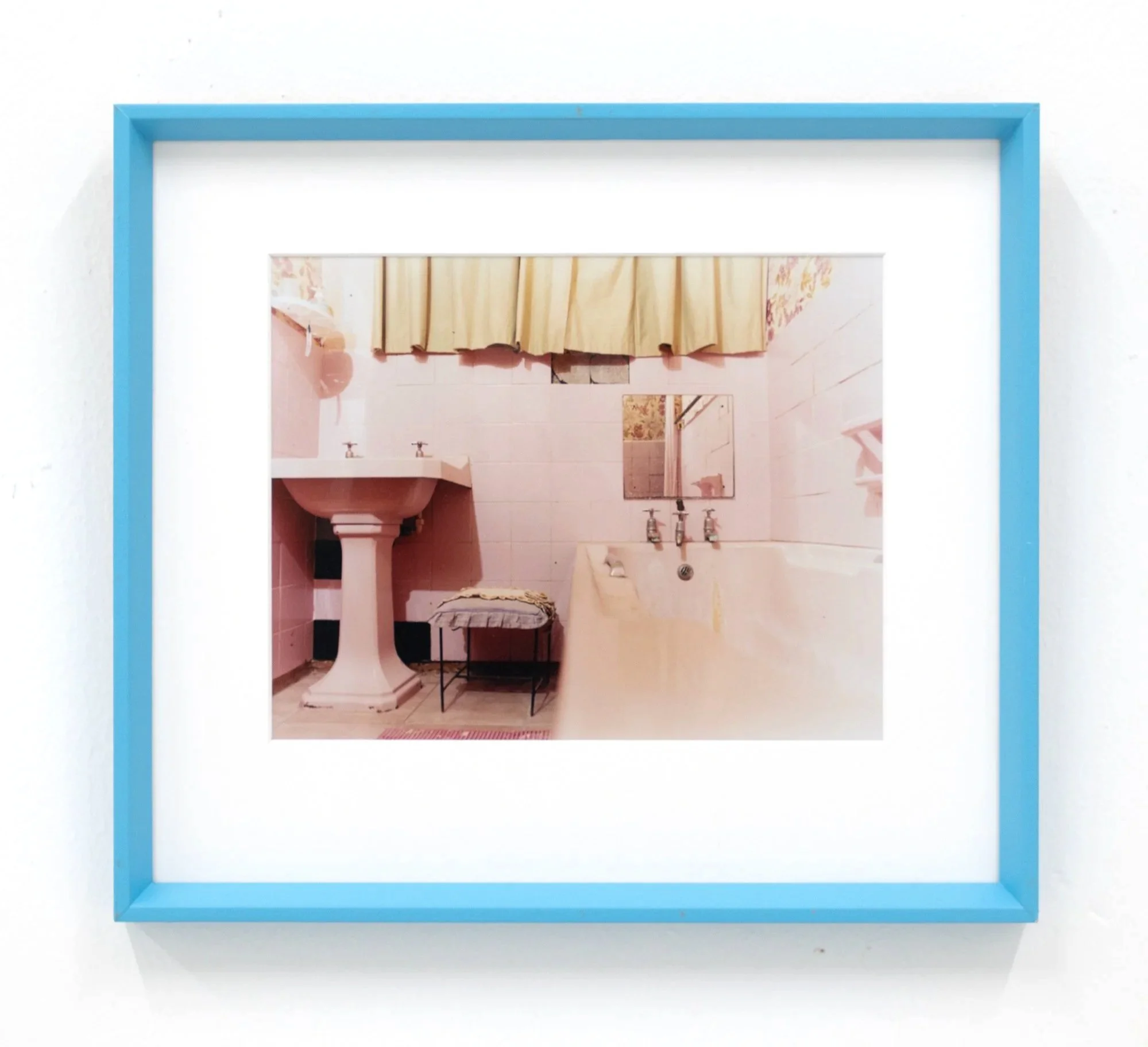 Photograph of a mirror with a blue frame, reflecting a bathroom with pink walls, a mirror, a sink, and a towel.