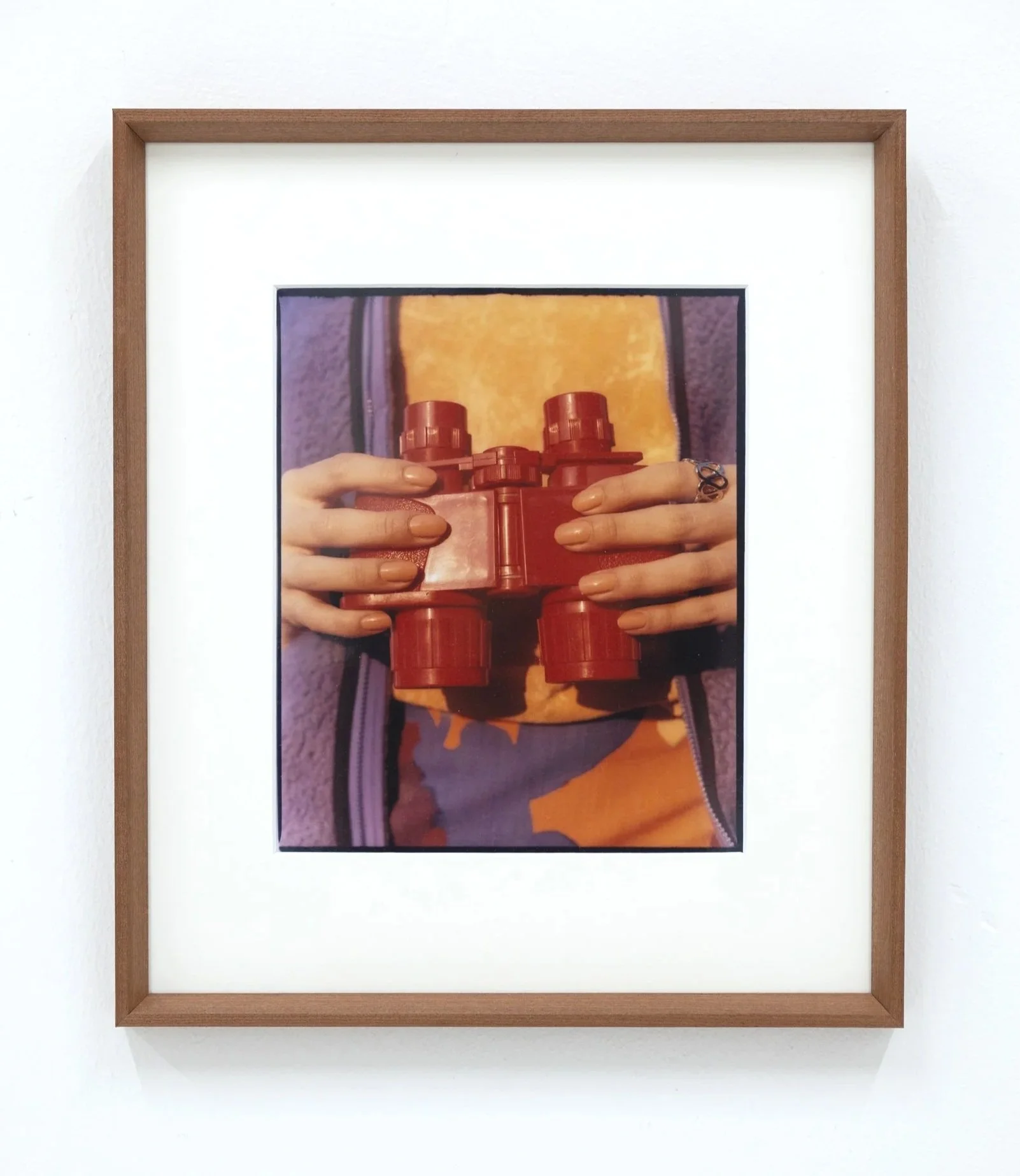 Framed photograph of a person holding a red object with both hands, wearing a ring and a yellow shirt with an orange design, against a purple background.