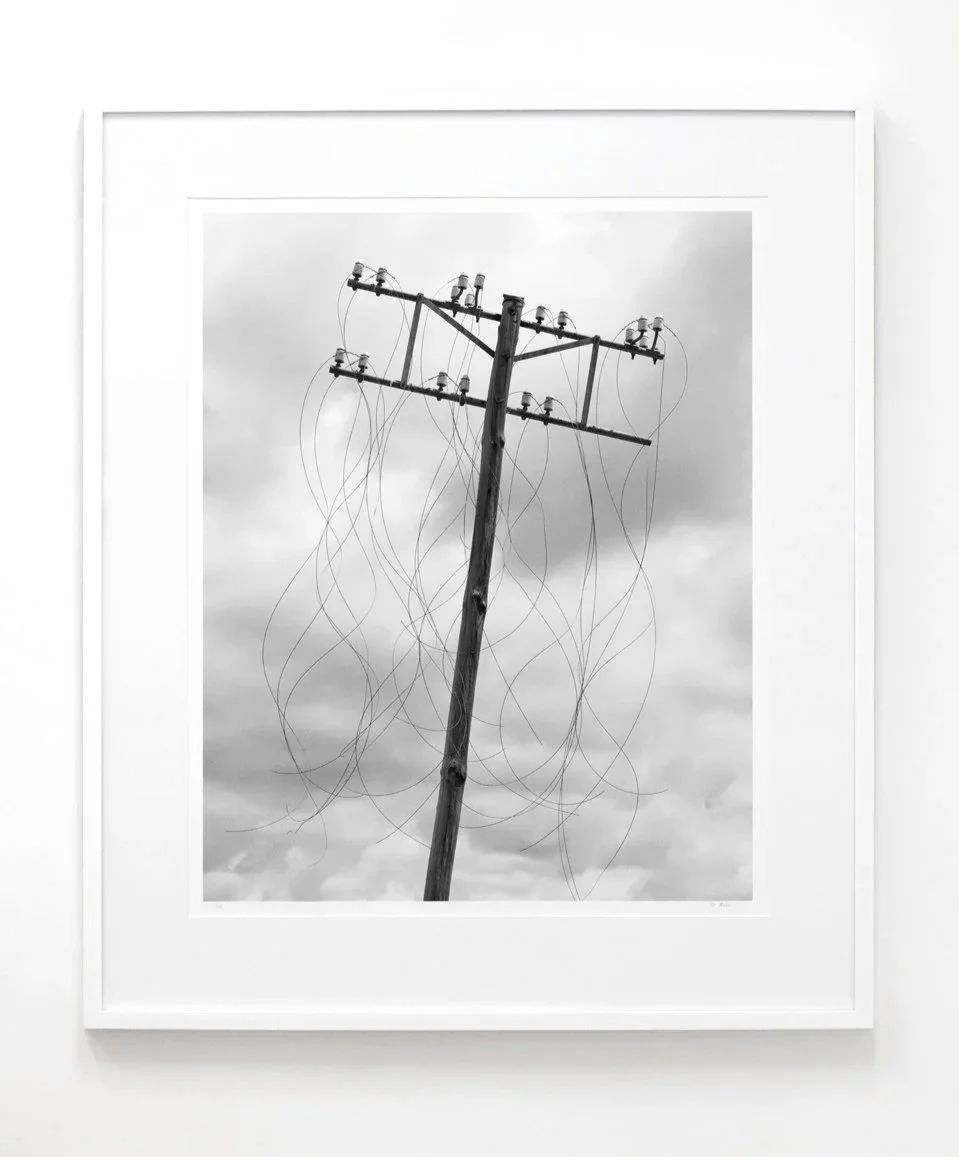Framed black and white photograph of an electrical pole with multiple wires against cloudy sky, framed and hung on a white wall.
