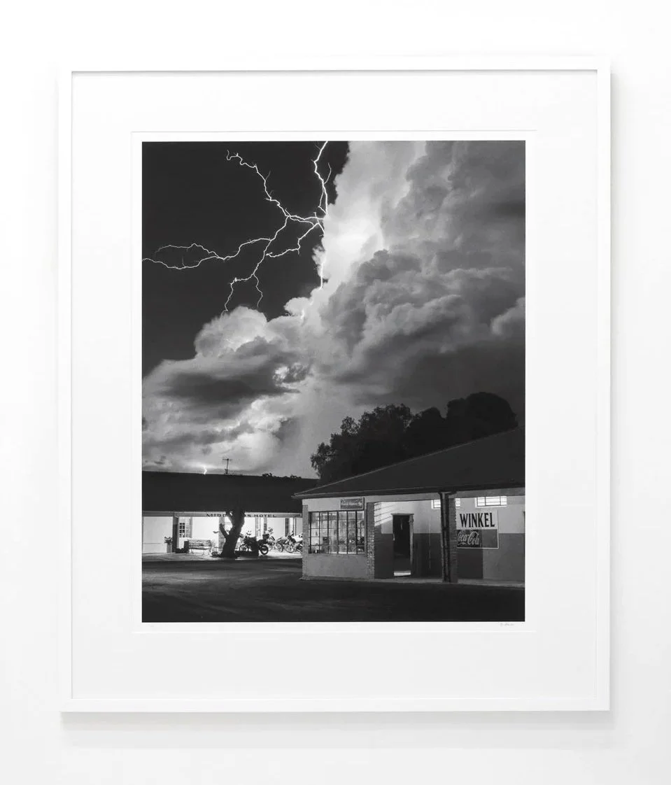 Framed black and white photograph of a sky with lightning and storm clouds over a small building and trees.