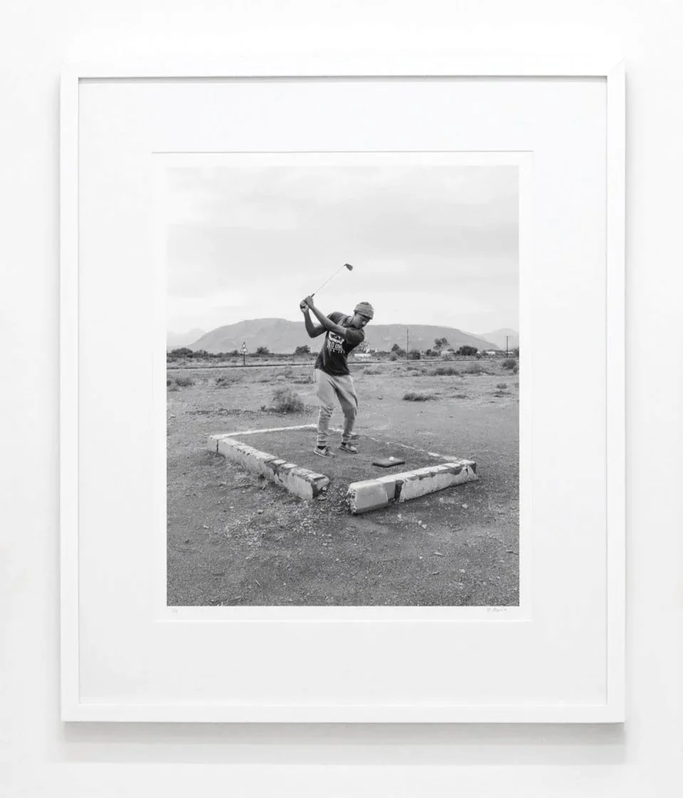 Black and white photograph of a man mid-swing on a golf course, in a desert landscape with mountains in the background, framed and hung on a white wall.