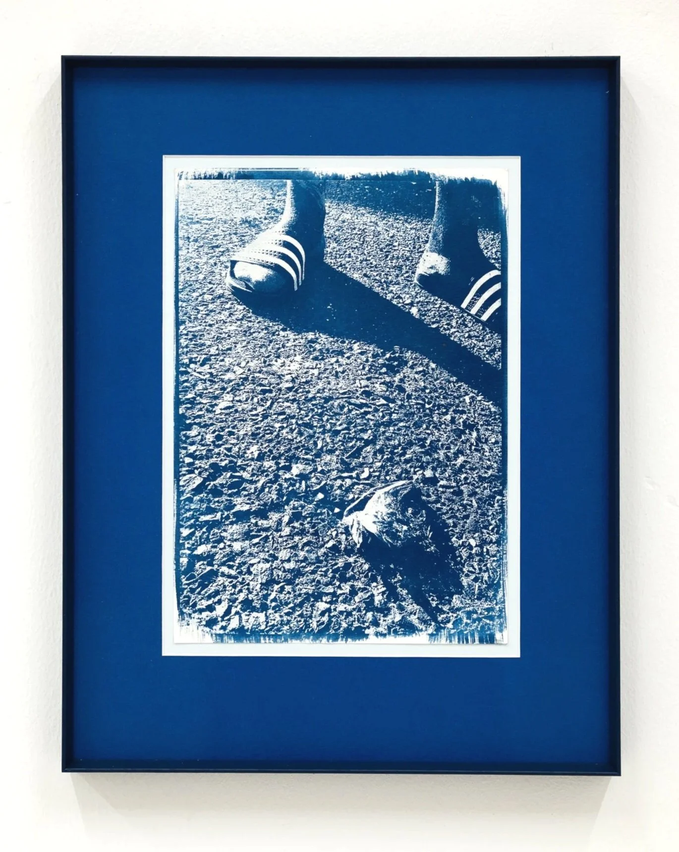 Black and white photograph of feet in sneakers on rocky ground, cast shadow and a stone in the foreground