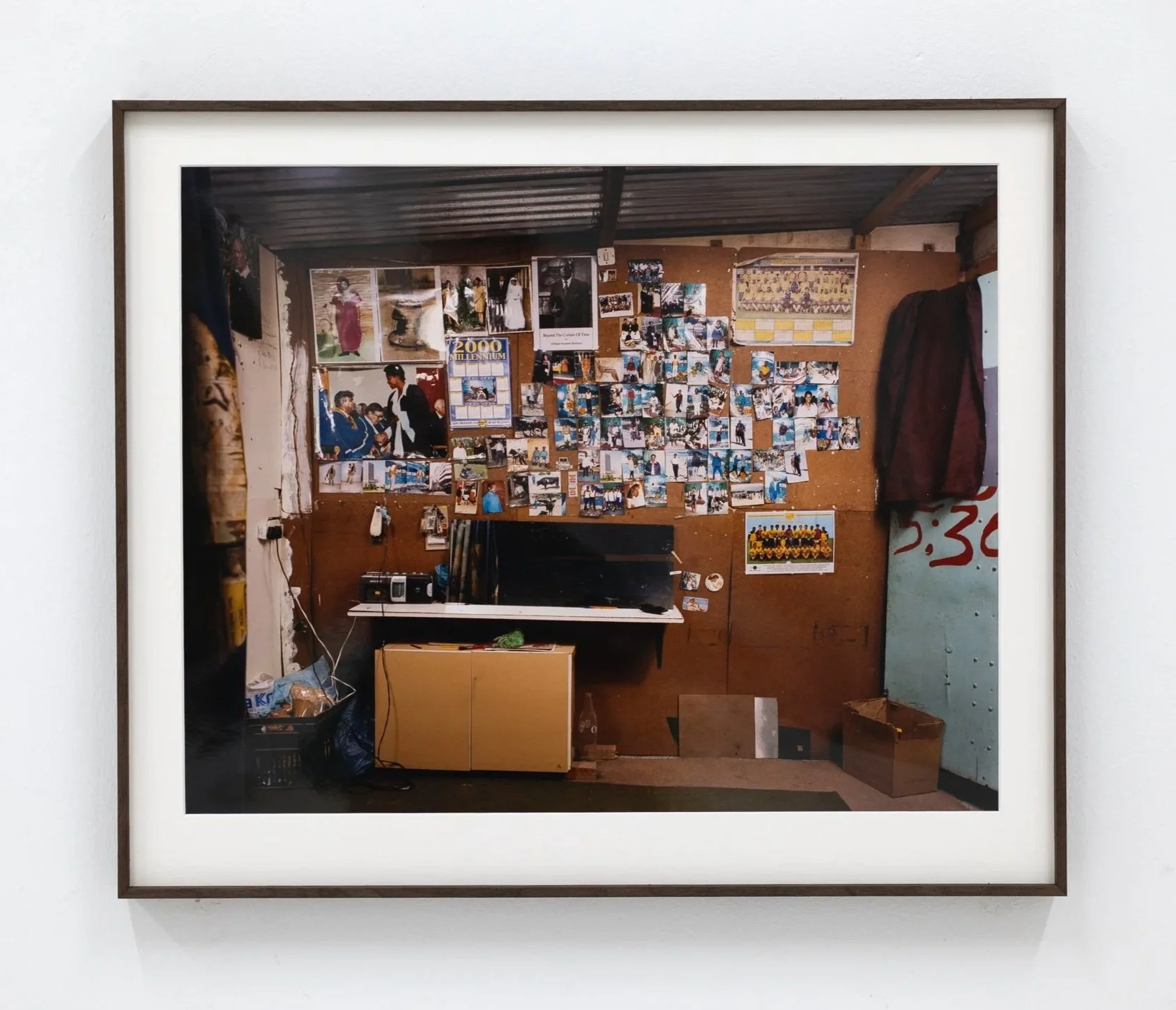 A framed photograph on a white wall showing a view of a cluttered room with bulletin boards, photographs, documents, a desk with a computer, and various personal items.