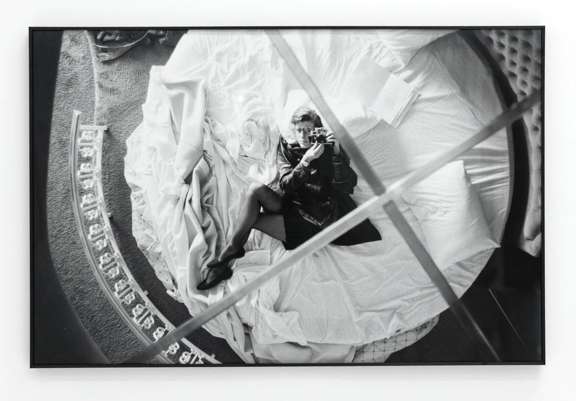Black and white photograph of a woman taking a selfie using a camera, reflected in a mirror on the floor, surrounded by crumpled bedsheets, books, and other items on a bed.