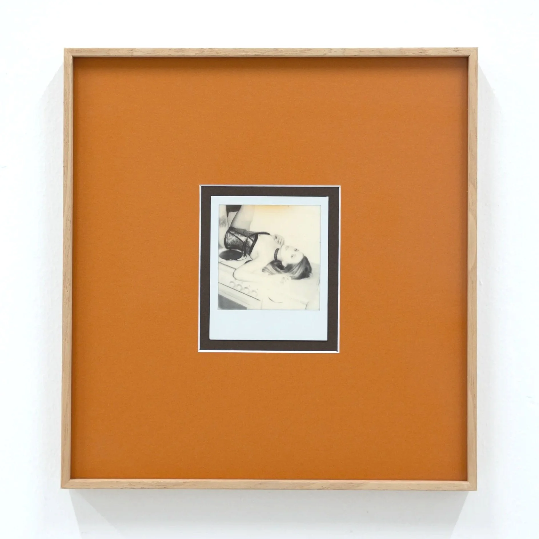 A framed black and white photograph of a woman lying on a bed, wearing lingerie and stockings, displayed inside an orange and wood frame on a white wall.