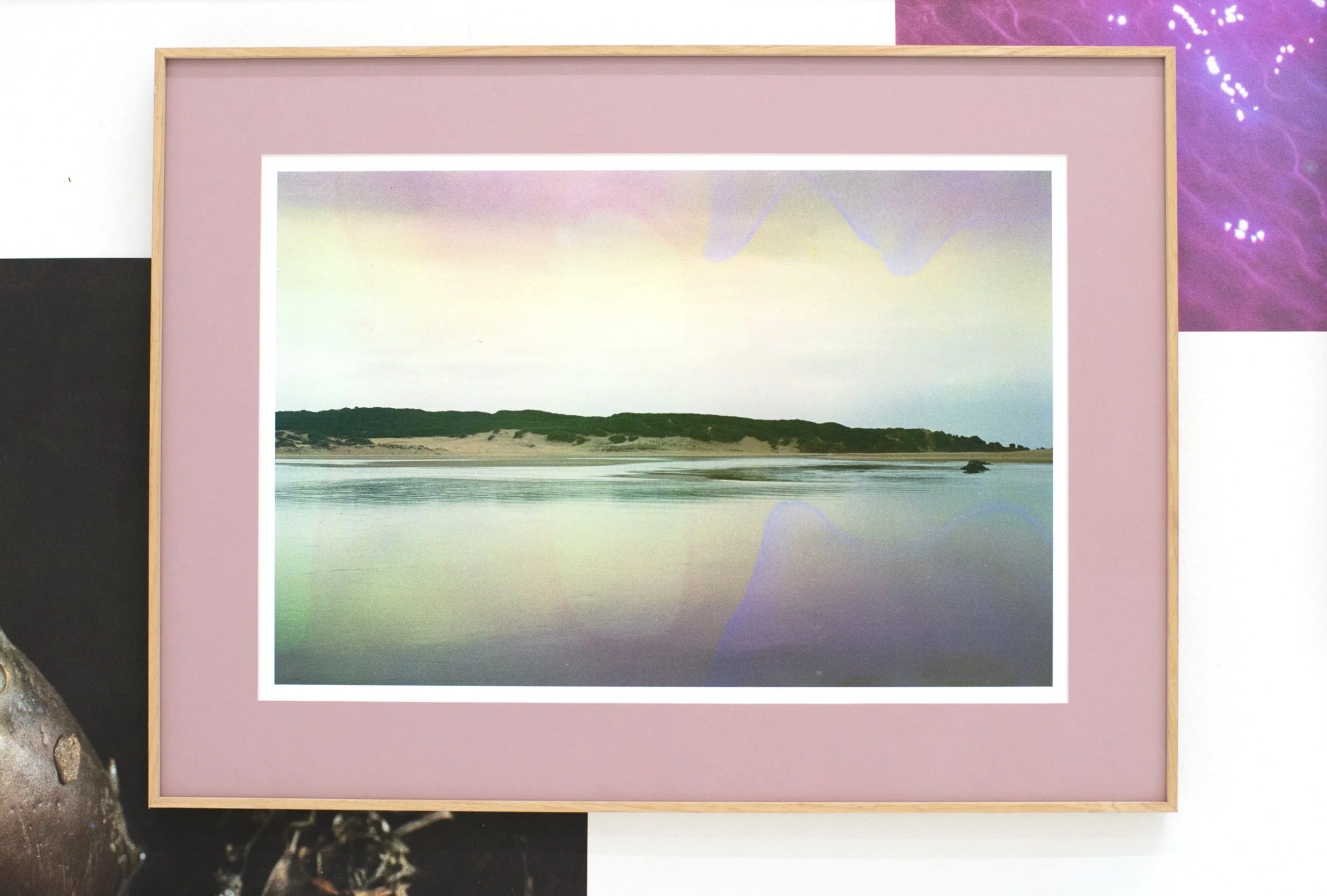 Photograph of a framed landscape photo featuring a beach with sand dunes and vegetation, set on a pink mat, against a multi-colored background.