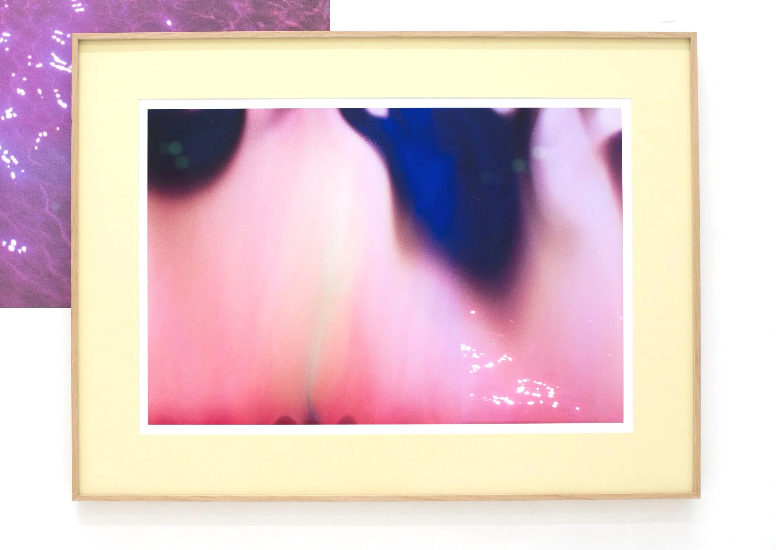 Close-up of an abstract art piece featuring a blurred face with pink and blue tones, framed with a light wooden border and mounted on a wall.