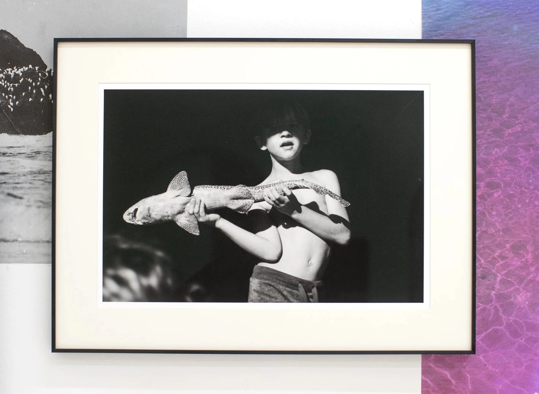 Black and white photograph of a boy holding a fish, posing shirtless against a dark background.