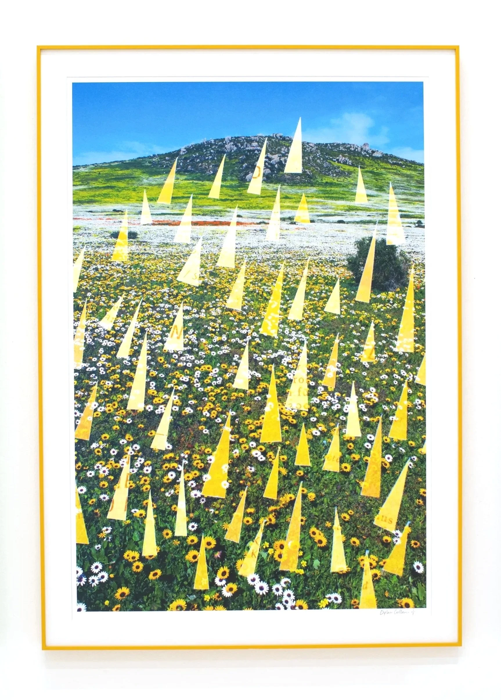 Colorful framed landscape photograph of a green hillside with a rocky hilltop against a bright blue sky, with yellow flags in the foreground.