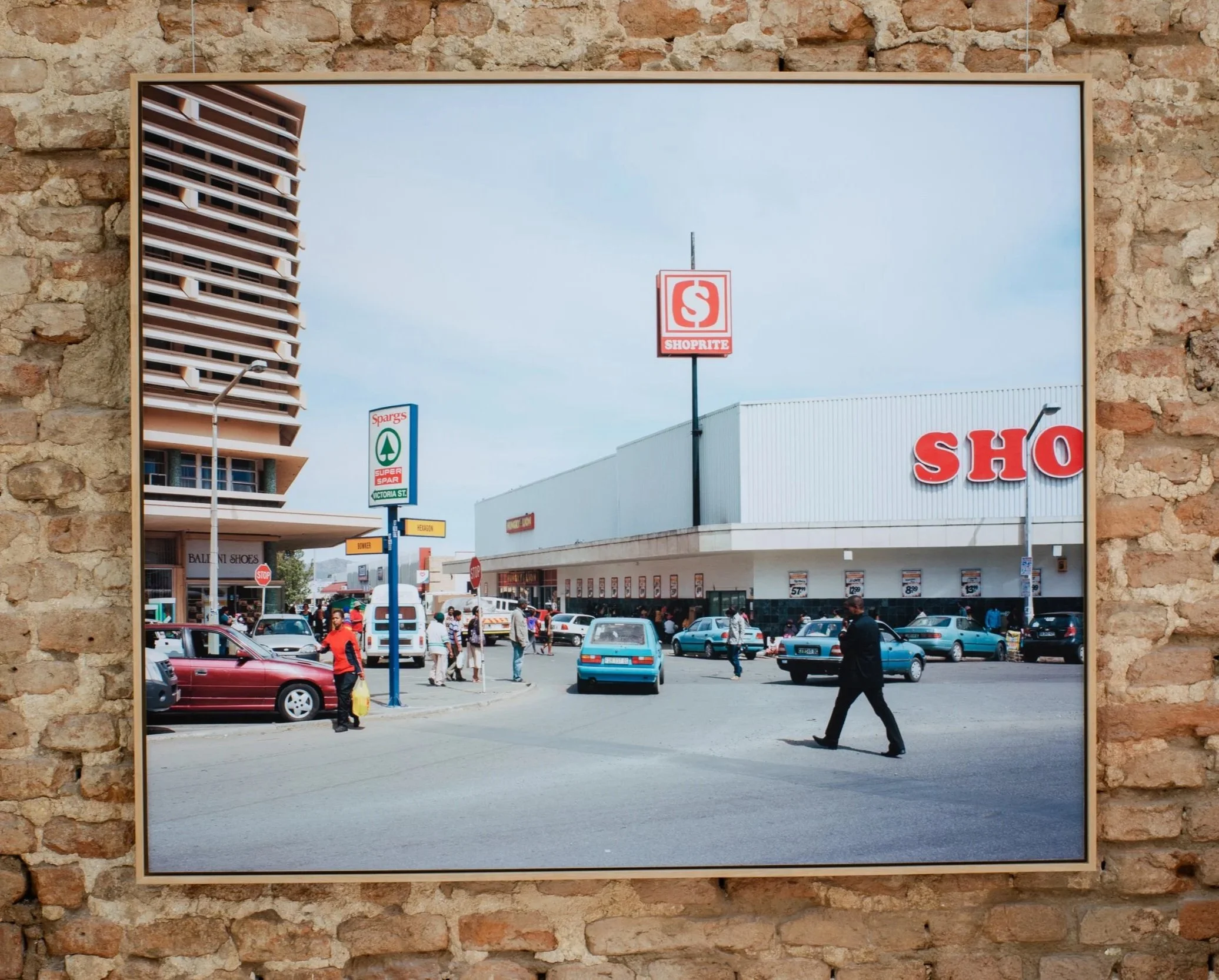 A parking lot in front of a shopping center with people walking, cars parked, and signs including Shoprite and Spargs, viewed through a mirror against a brick wall.