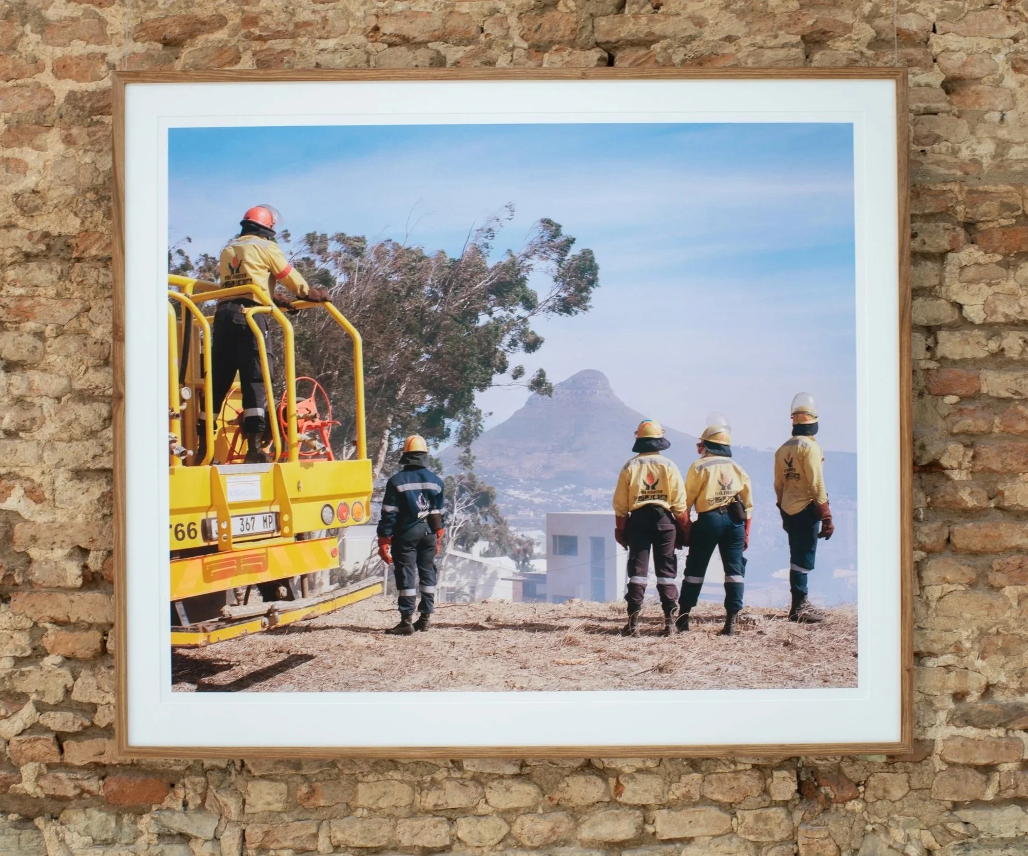 Framed photograph of firefighters working outdoors near a large tree and a mountain, displayed on a brick wall.