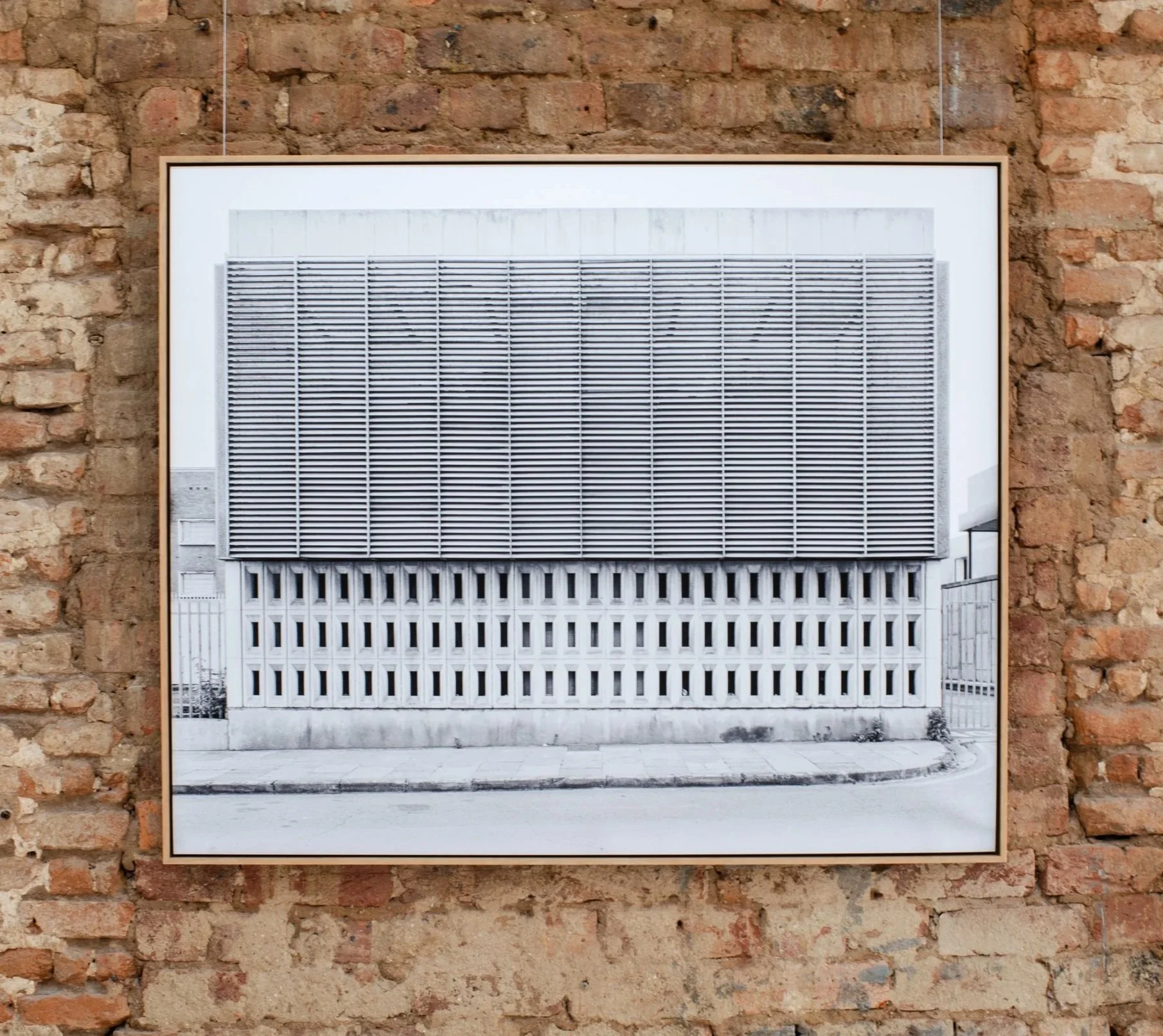 Photograph of a framed black and white photo of a building with horizontal lines, hung on a brick wall.