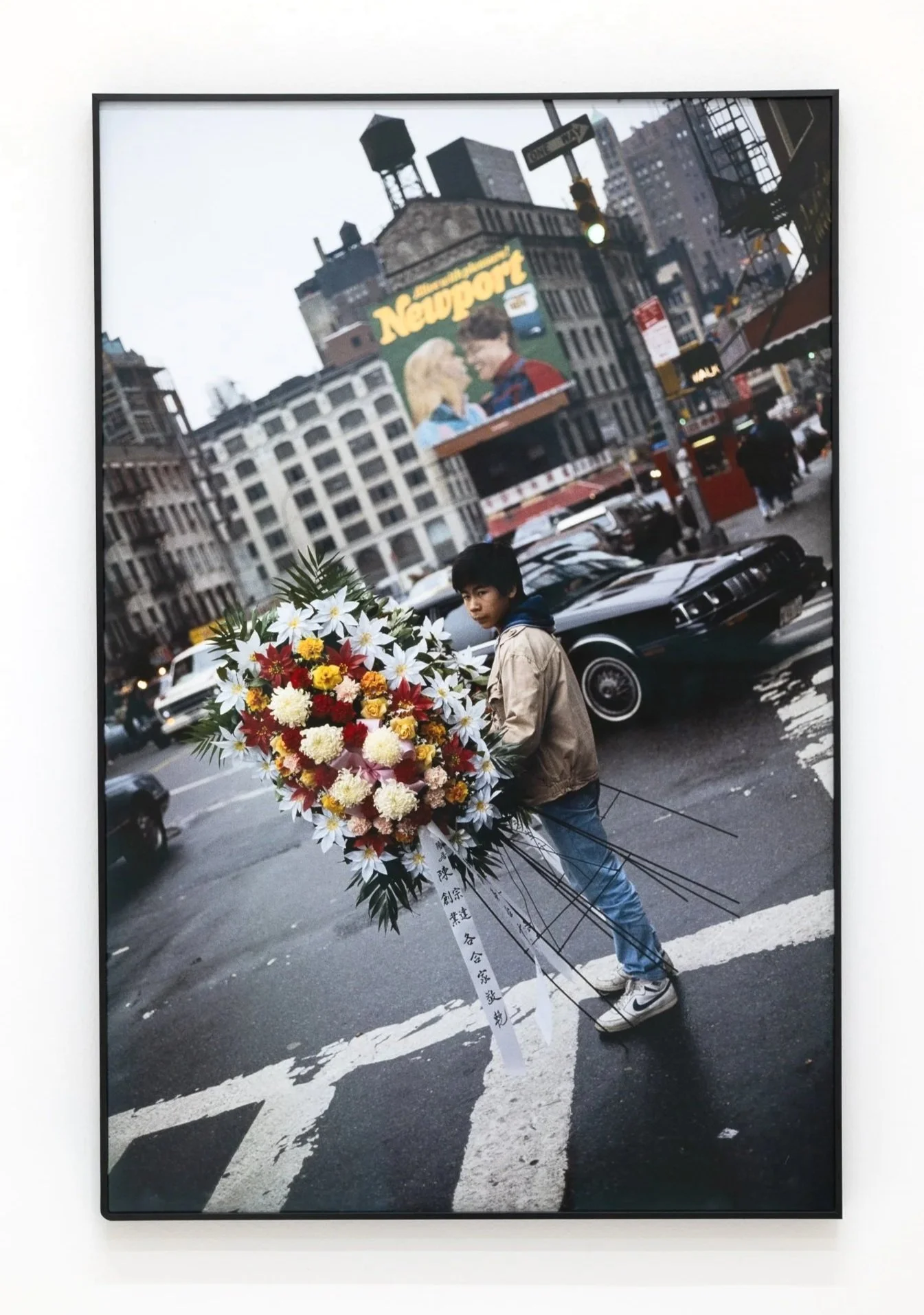 A young man holding a large flower arrangement with white, yellow, and red flowers, standing in the middle of a city street at a crosswalk, with cars in the background and buildings around him.