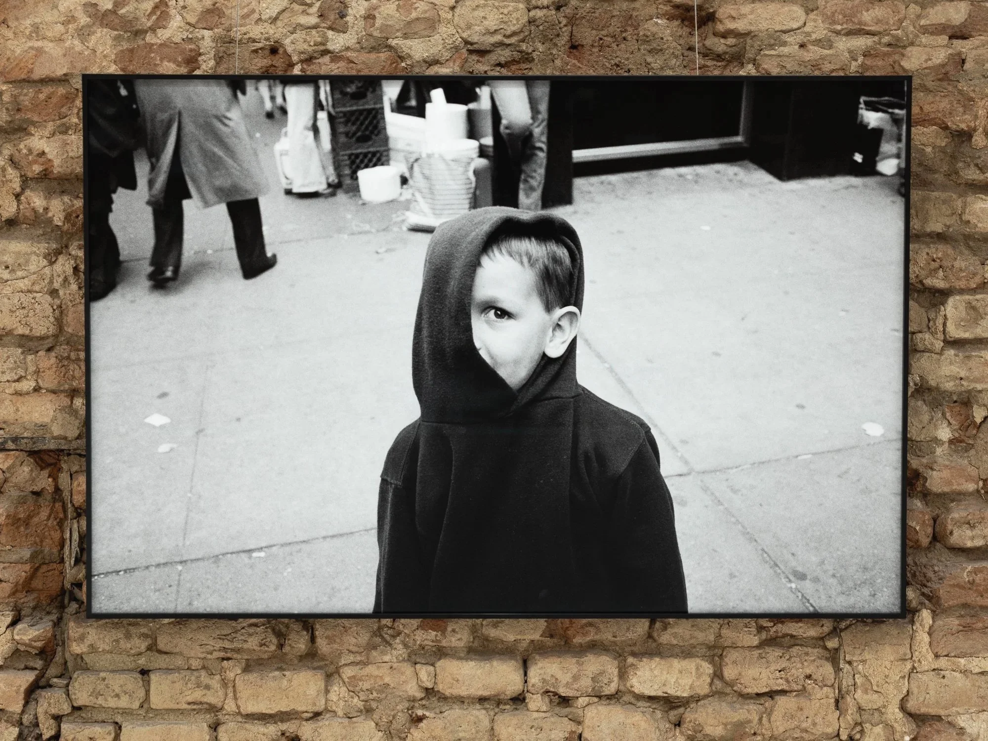 Framed black and white photograph of a boy with a hoodie, partially covering his face, standing outdoors on a concrete sidewalk.