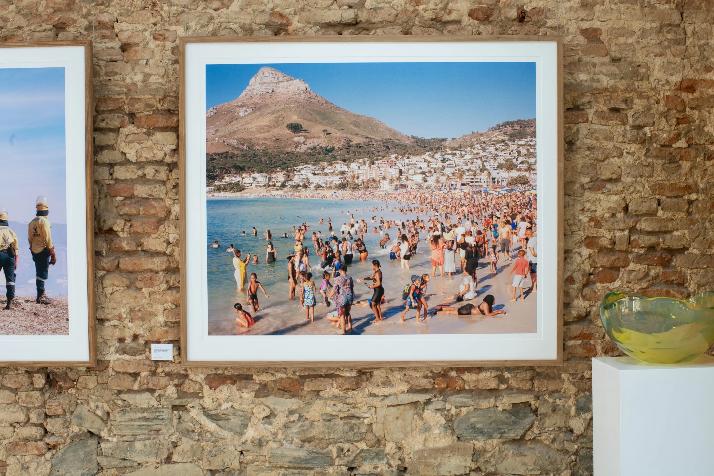 A framed photograph of a crowded beach scene with a mountain in the background, mounted on a rustic brick wall.